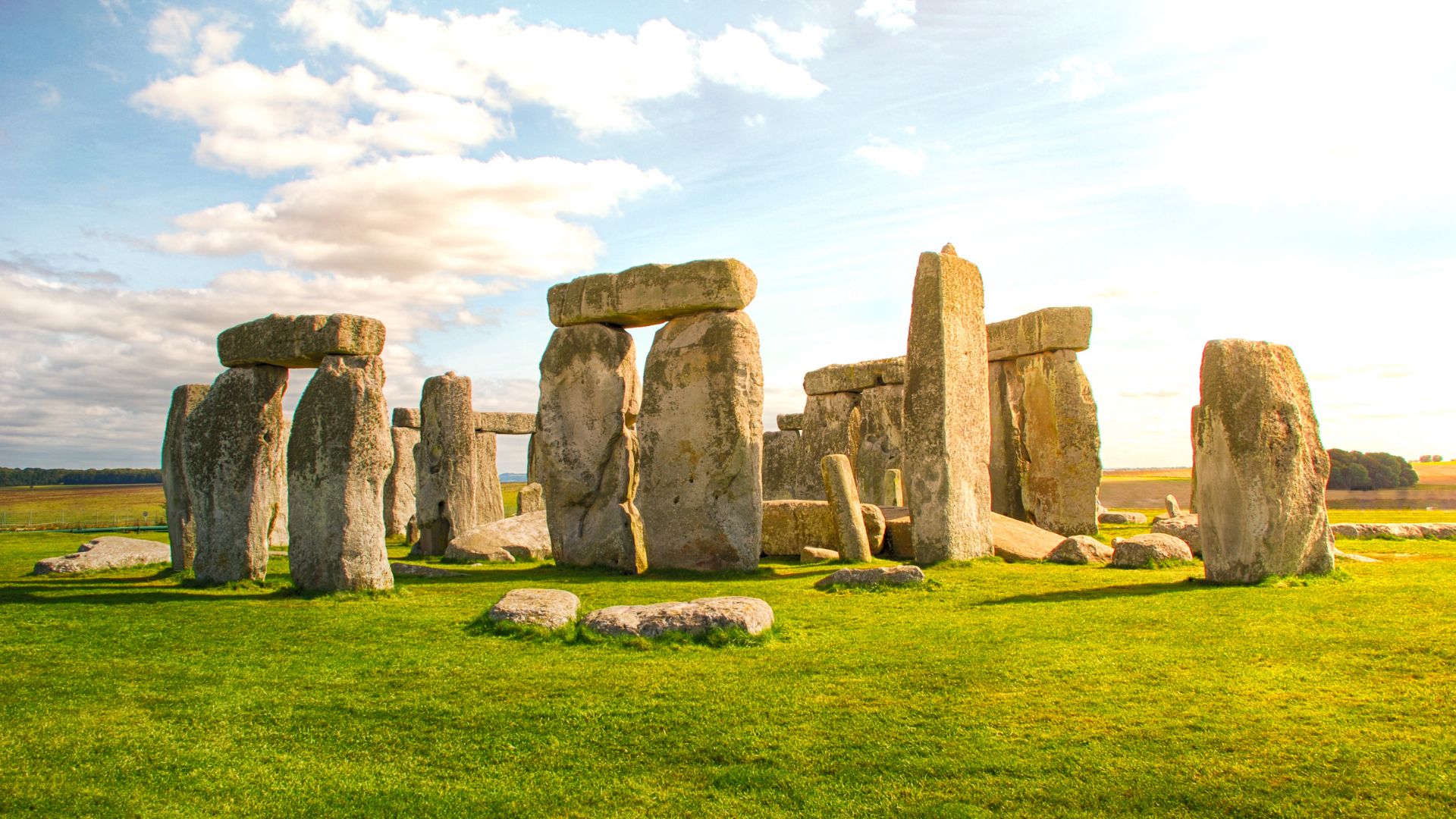A wide shot of Stonehenge, a prehistoric monument consisting of large standing stones arranged in a circular pattern, set against a green grassy plain under a partly cloudy sky.