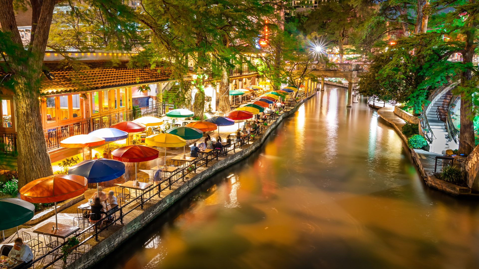 A vibrant evening scene of the San Antonio River Walk, featuring a tree-lined river with calm water, and a pedestrian promenade on the left lined with numerous outdoor dining areas under colorful umbrellas, illuminated by warm lights.