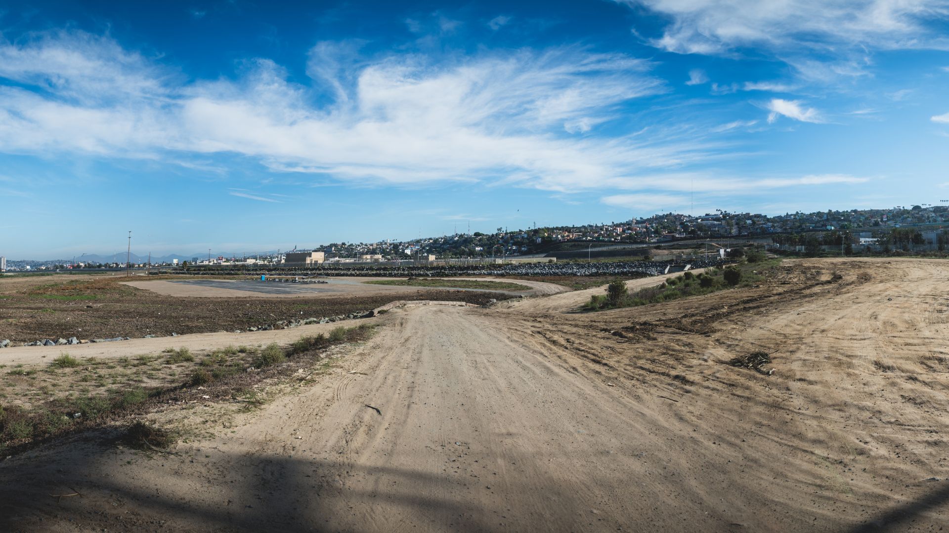 A wide-angle view of a dry, unpaved area leading towards a distant urban landscape under a blue sky with scattered clouds, indicating the US-Mexico border near San Diego and Tijuana. 