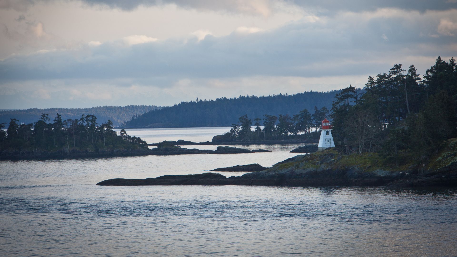 A wide shot of a body of water with several tree-covered islands and rocky outcrops under a cloudy sky. A small white lighthouse with a red top stands on a rocky island in the middle ground on the right side of the frame.