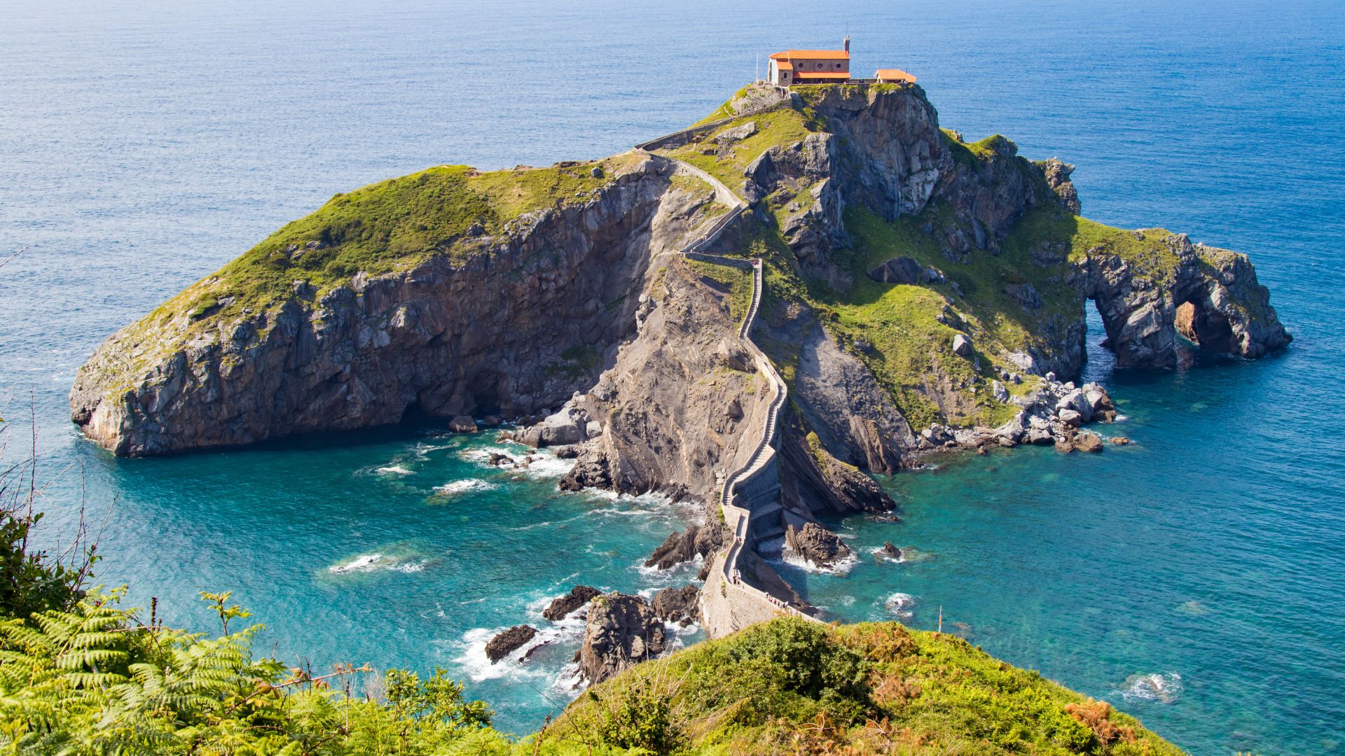 A high-angle shot captures the iconic San Juan de Gaztelugatxe, an island hermitage connected to the mainland by a winding stone bridge and a long, steep staircase, all set against the vibrant blue waters of the Cantabrian Sea and surrounded by green cliffs.
