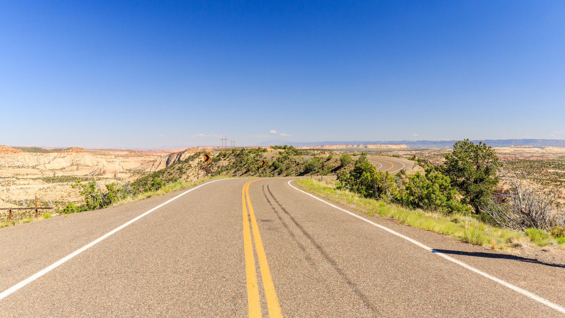 A winding, two-lane paved road with a double yellow line ascends a ridge in a desert landscape under a clear blue sky, with distant mesas visible on either side.