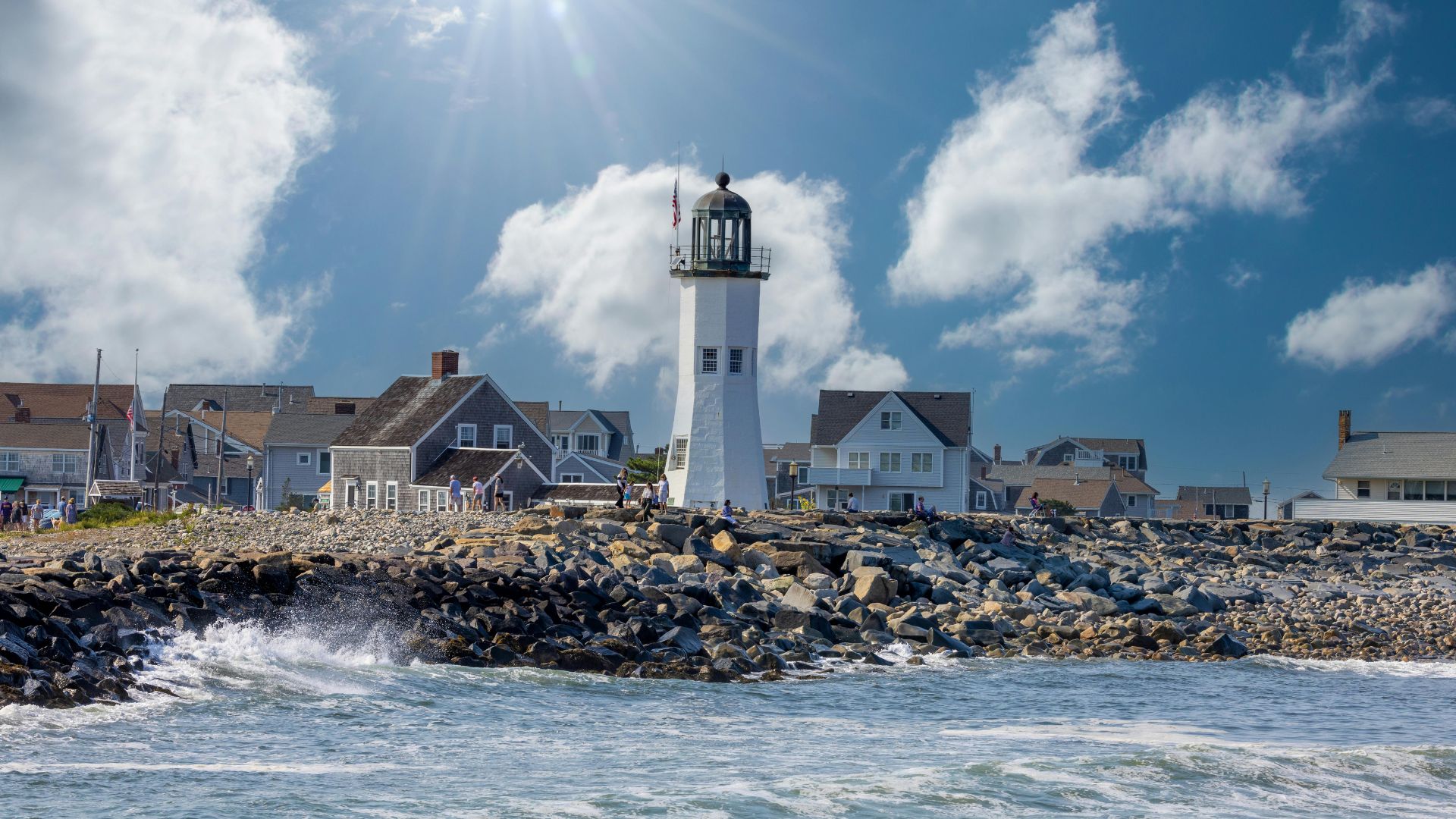 A white lighthouse stands on a rocky shore next to the ocean, with waves crashing in the foreground and houses visible behind the lighthouse under a partly cloudy sky.