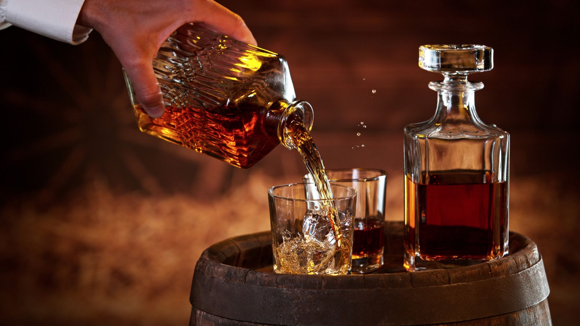 A hand pours amber-colored whisky from a square bottle into a glass, with a full decanter and another glass resting on a wooden barrel.