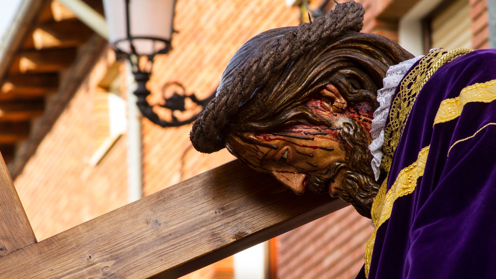  A close-up shot of a religious sculpture depicting Jesus Christ, with a pained expression and bloodied face, carrying a wooden cross on his shoulder, dressed in a rich purple and gold robe during a Holy Week procession in Spain. 