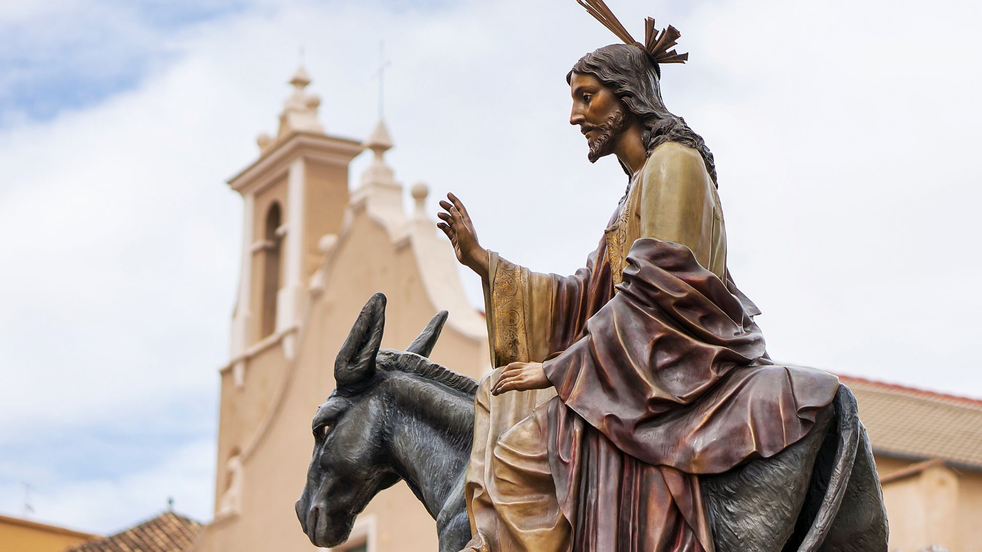 A statue of Jesus Christ riding a donkey during a Holy Week procession in Spain, with a church building visible in the background.