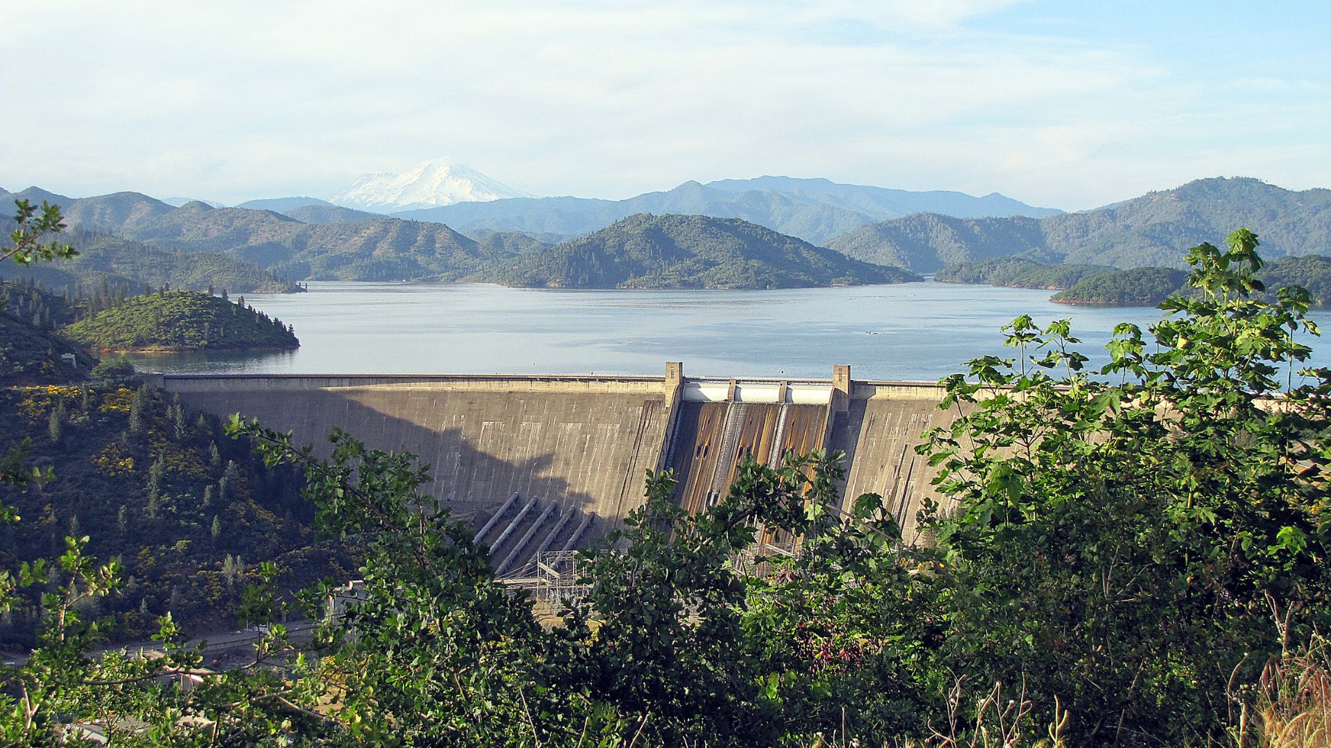 A wide shot of Shasta Dam with Shasta Lake in the background, surrounded by green, tree-covered mountains, and a snow-capped peak visible in the distance. The dam is a large concrete structure with several spillway gates, and in the foreground, lush green trees frame the bottom of the image. 