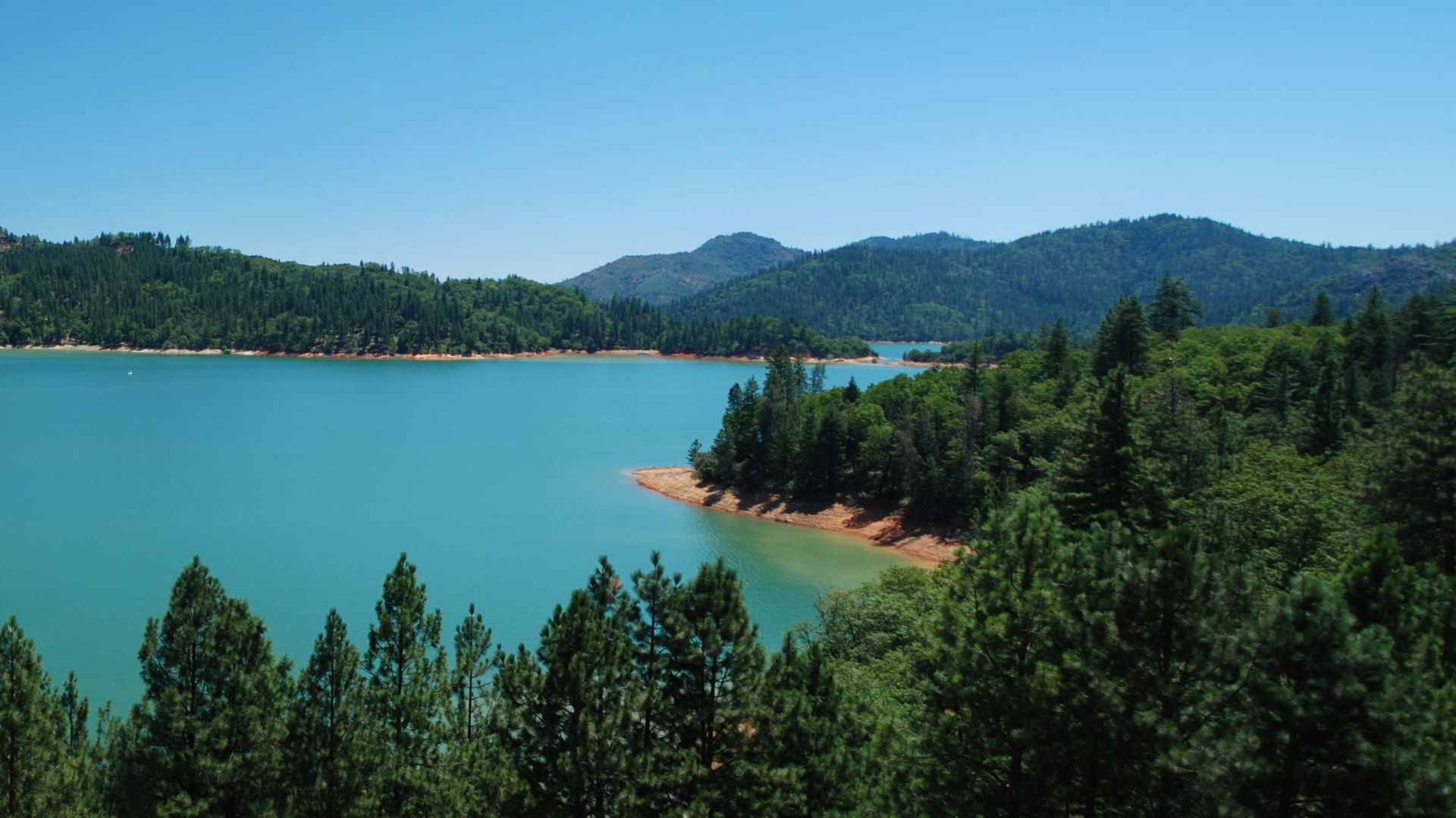 A wide shot of a large, turquoise-blue lake surrounded by dense green forests and rolling mountains under a clear blue sky.