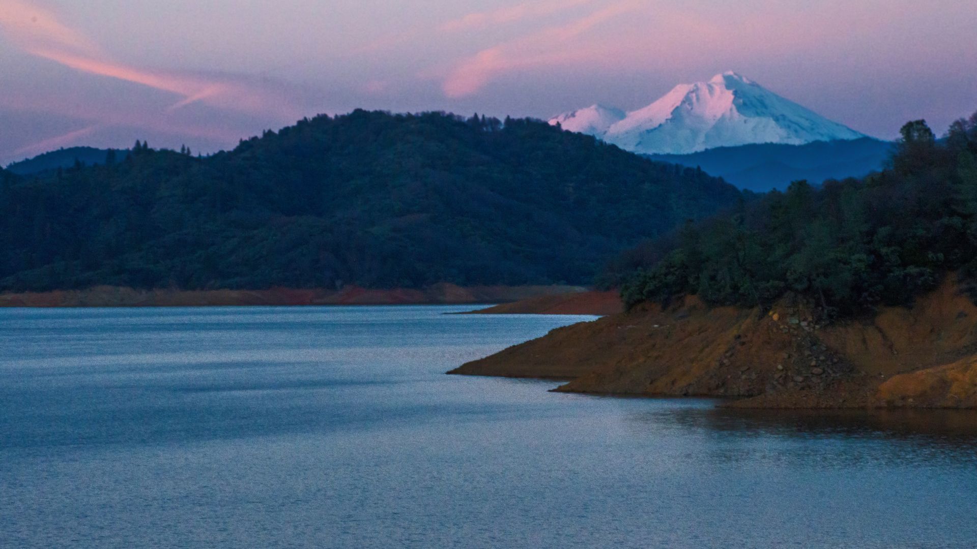 A wide shot of Shasta Lake at dusk, with dark hills surrounding the lake and snow-capped Mount Shasta visible in the distance under a colorful sky.