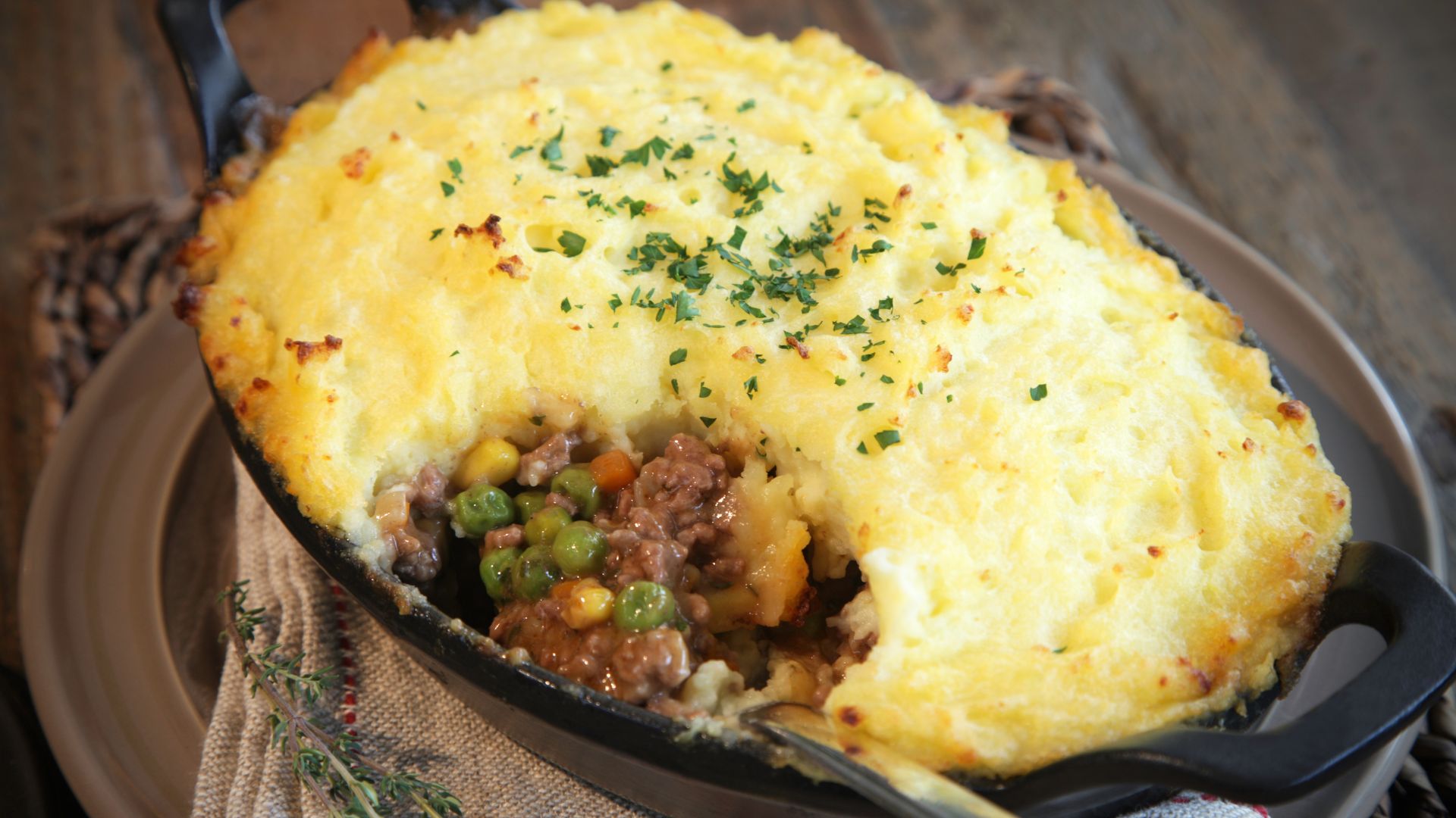 A close-up of a Shepherd's pie in a black baking dish, showing a scoop removed to reveal the meat and vegetable filling (peas, corn, carrots) beneath a golden-brown mashed potato topping, garnished with chopped herbs.