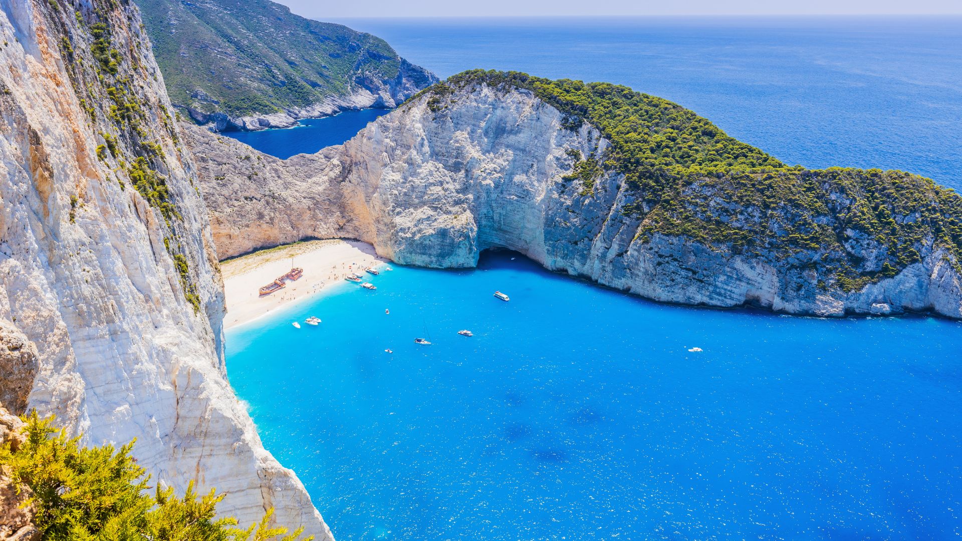 An aerial view of Navagio Beach on Zakynthos, Greece, showcasing its striking turquoise waters, white pebble beach, and dramatic white cliffs surrounding the cove, with several small boats visible in the water.