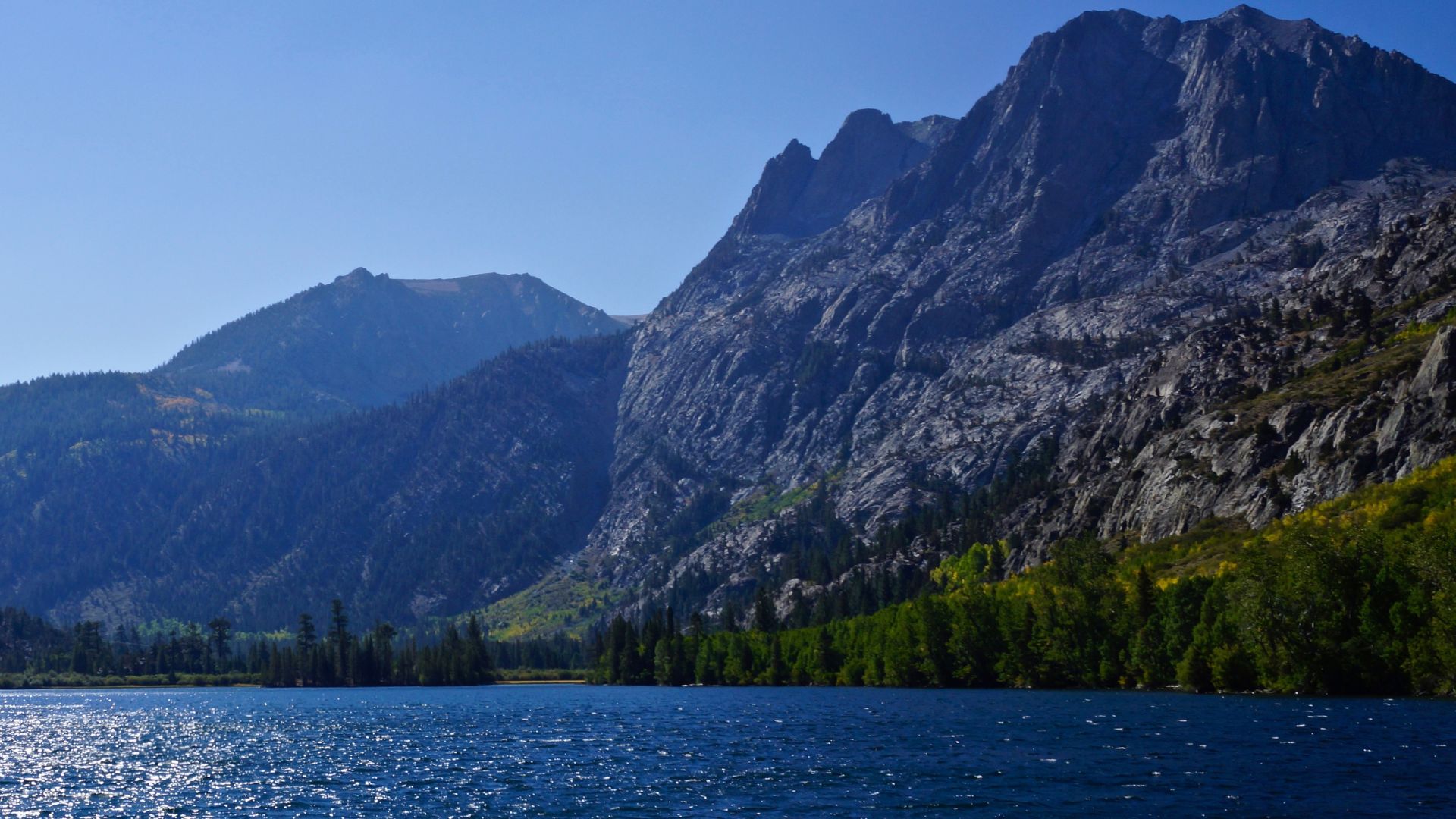 A serene blue lake reflecting a clear sky, surrounded by lush green trees and towering, rugged mountains under a bright, sunny day.