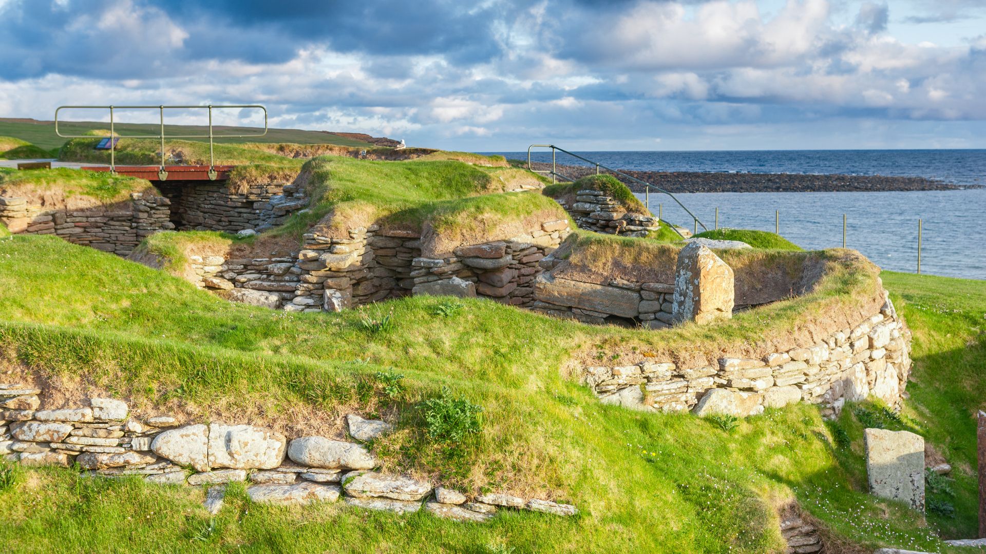A panoramic view of Skara Brae, a well-preserved Neolithic settlement on the Orkney Islands, featuring ancient stone dwellings with grassy roofs built into a coastal landscape, with the ocean visible in the background under a partly cloudy sky.