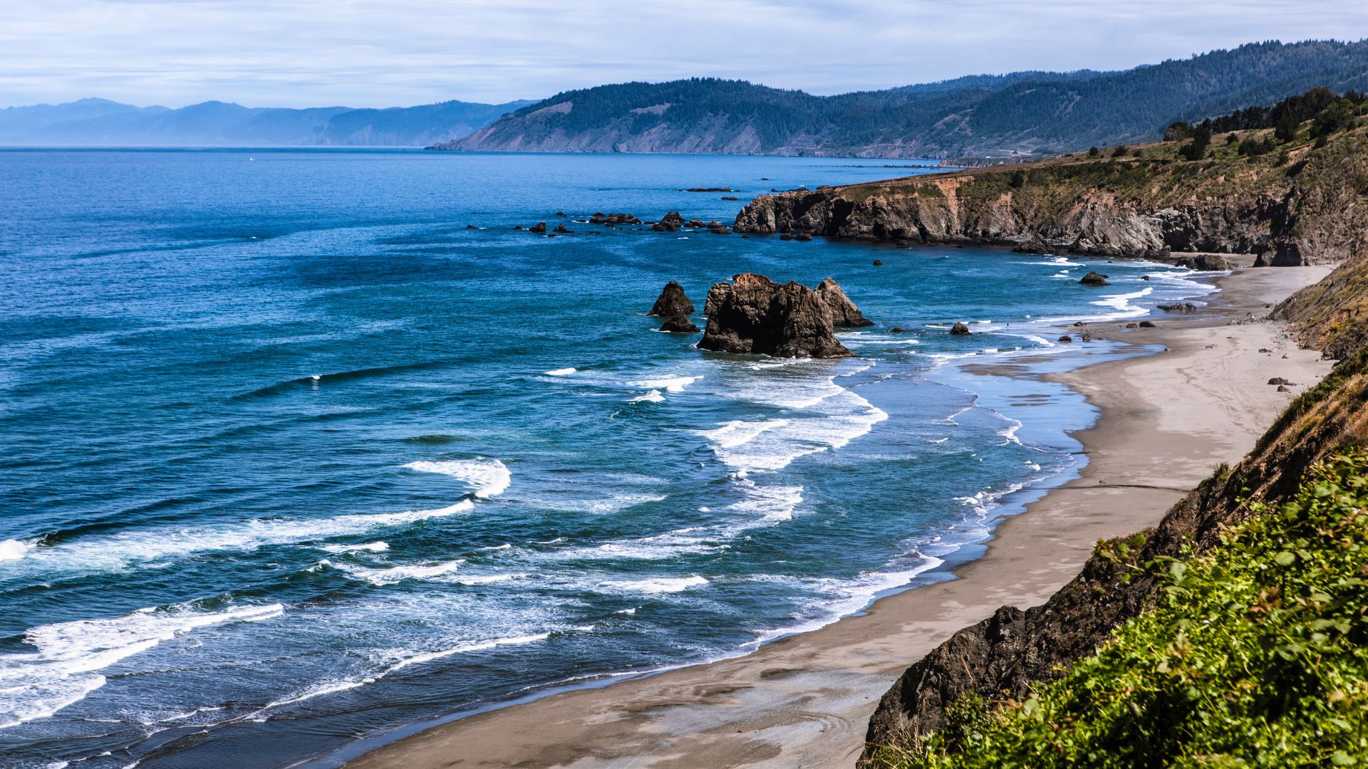 A sweeping view of a rugged coastline with a sandy beach meeting the Pacific Ocean, featuring large rock formations in the water and cliffs covered in green vegetation rising on the right, under a partly cloudy sky.