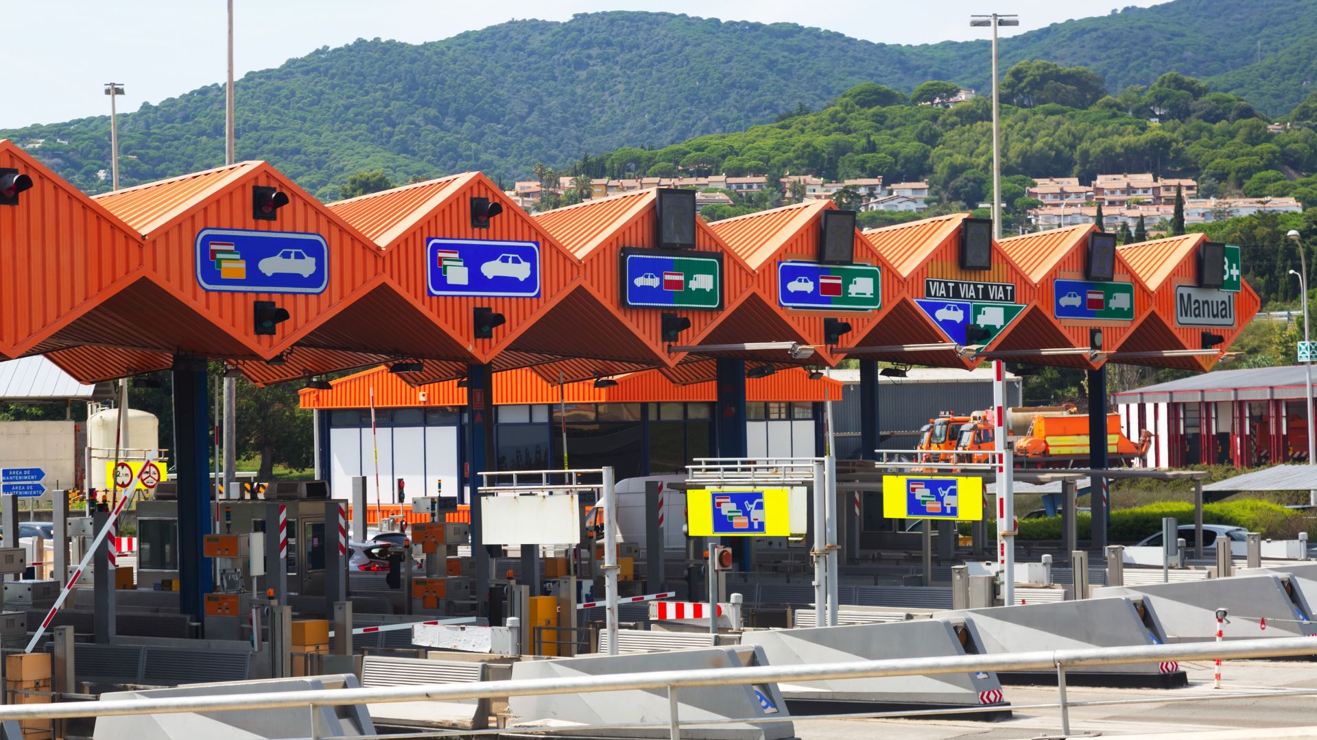 A wide shot of a multi-lane toll plaza featuring orange-roofed booths and electronic signage under a clear sky, with mountains visible in the background. Signs indicate different payment methods, including "Manual". 