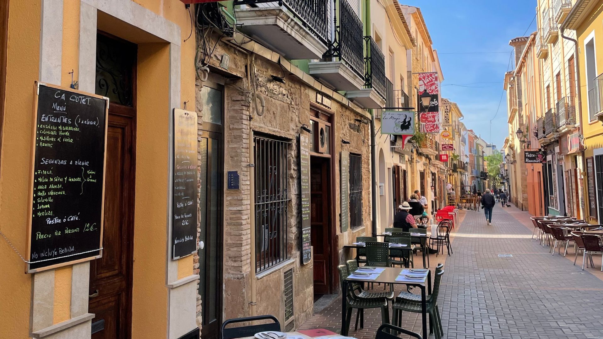 A narrow, pedestrianized street in Spain, lined with historic buildings featuring balconies and awnings. The street is bustling with outdoor seating areas for restaurants, with tables and chairs extending onto the cobblestone path. A menu board is visible on the left, and people are seen walking and dining along the street, under a clear blue sky. 