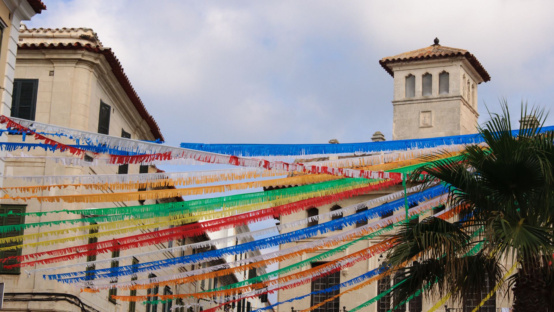 Colorful streamers adorn a street in Mahón, Menorca, Spain, with traditional buildings and a prominent bell tower under a cloudy sky, likely during a local fiesta.