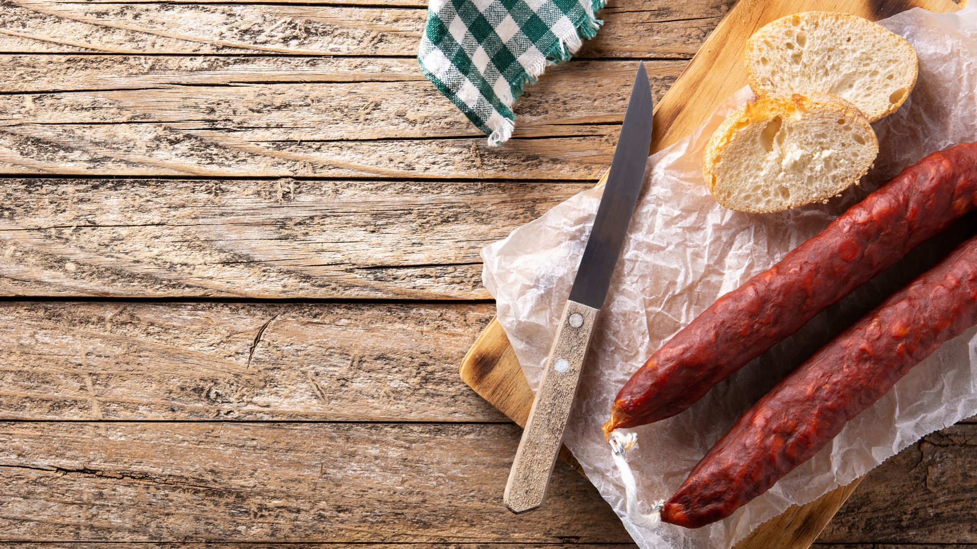 A close-up of two cured sausages, Spanish chorizo, alongside sliced bread and a knife on a wooden cutting board, all set on a rustic wooden table.