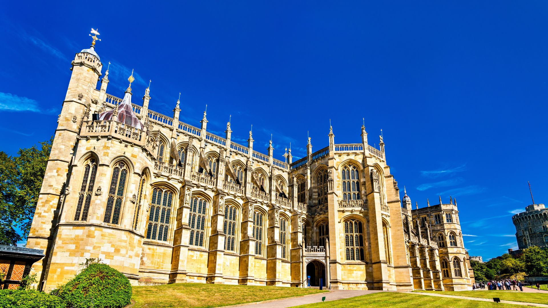 A wide exterior view of St George's Chapel at Windsor Castle under a bright blue sky, showcasing its Gothic architecture with tall arched windows, intricate stone details, and spires. 