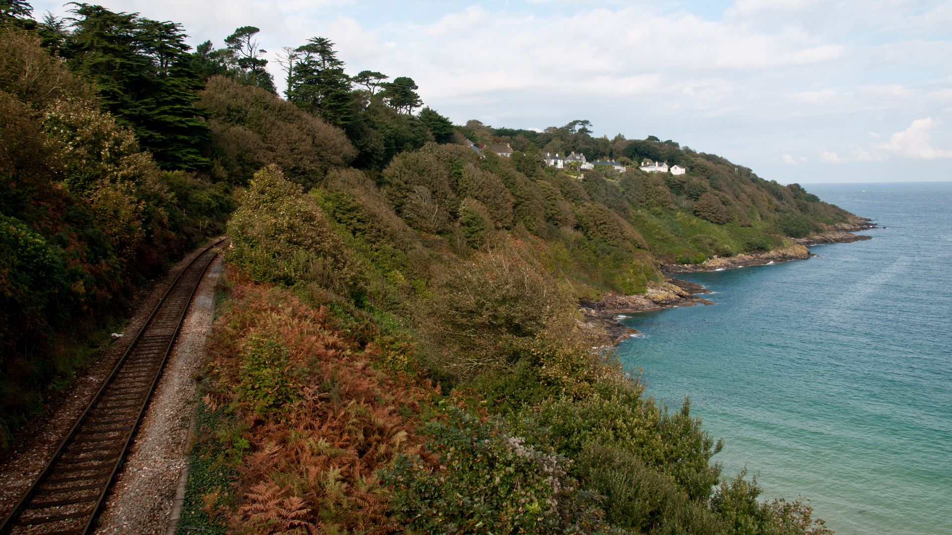 A train track runs along a steep, tree-covered coastline next to the turquoise sea under a cloudy sky, with houses visible on the distant clifftop.
