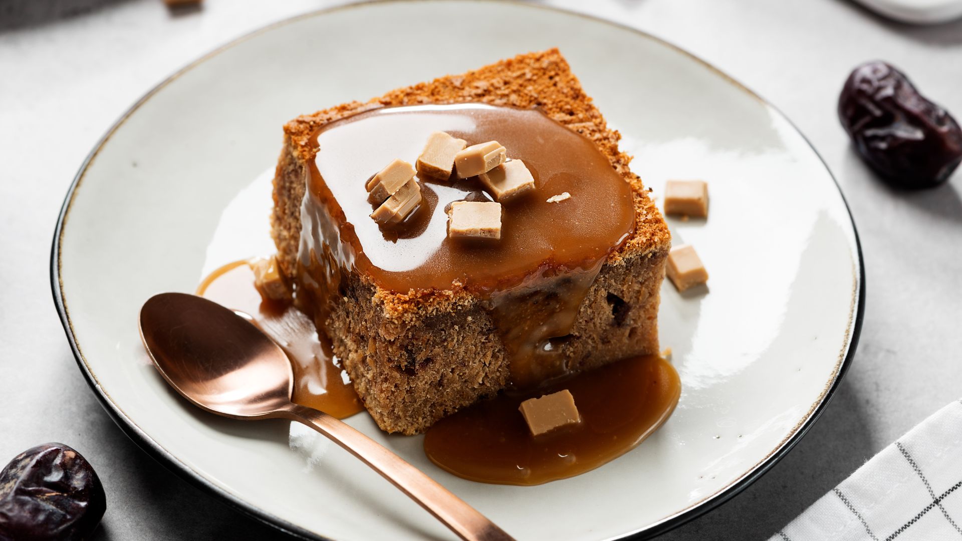 A slice of sticky toffee pudding with a rich toffee sauce and scattered toffee candy pieces on top, served on a light-colored plate with a copper spoon beside it.