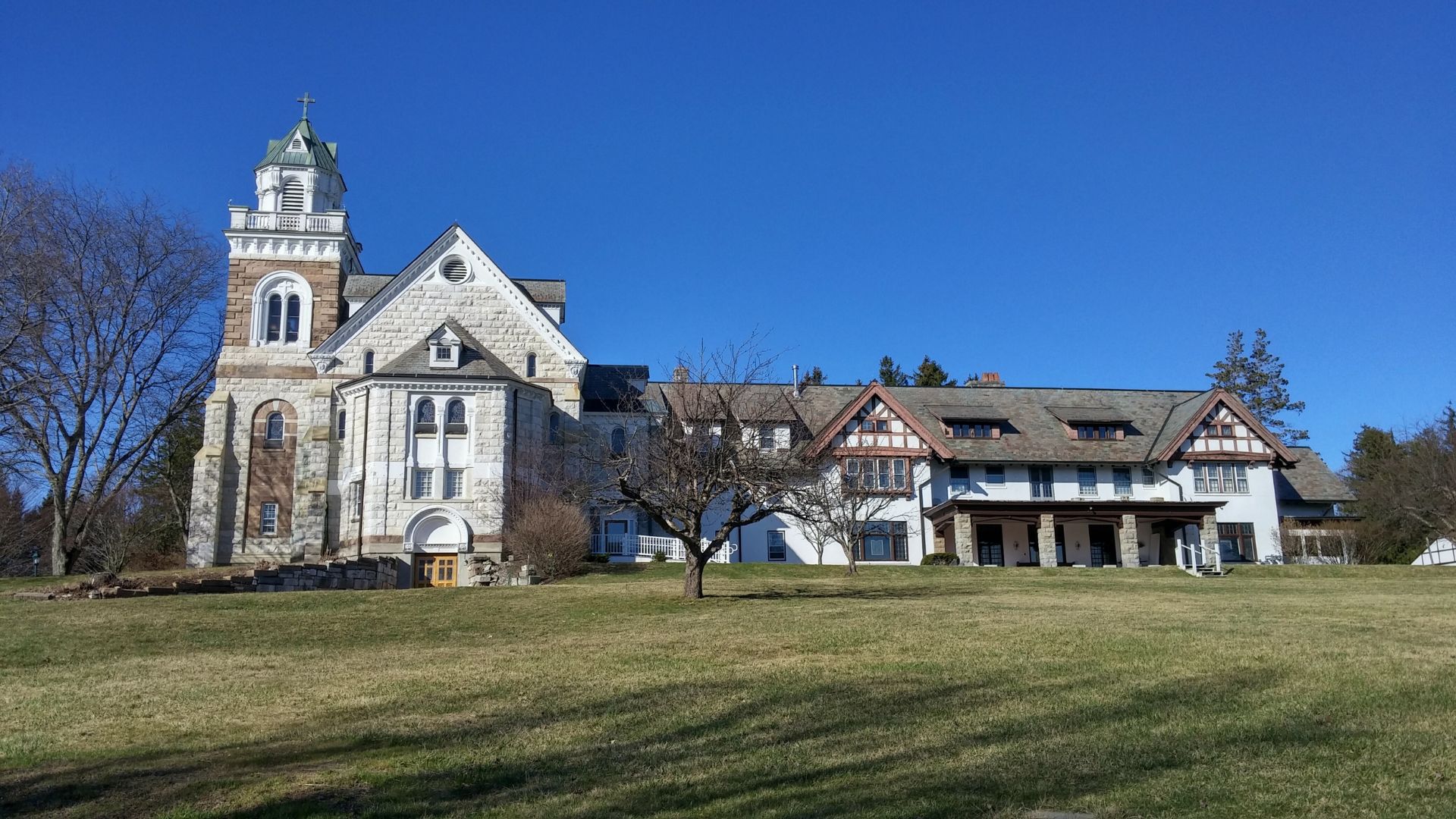 A wide shot of the National Shrine of The Divine Mercy, a large stone church with a prominent tower, and an attached multi-story building with a gabled roof, situated on a grassy hill under a clear blue sky.