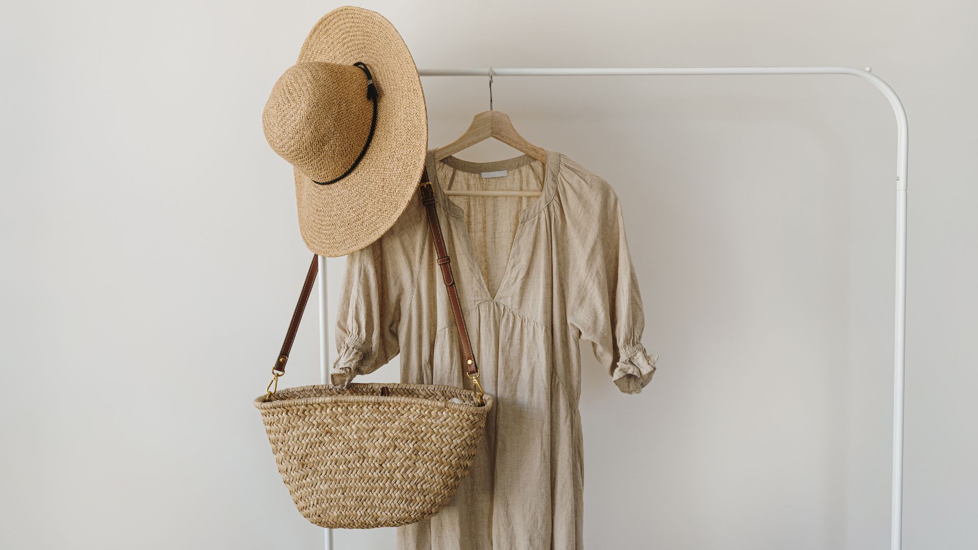 A beige dress hanging on a white rack, with a straw hat and a woven straw bag hanging next to it.