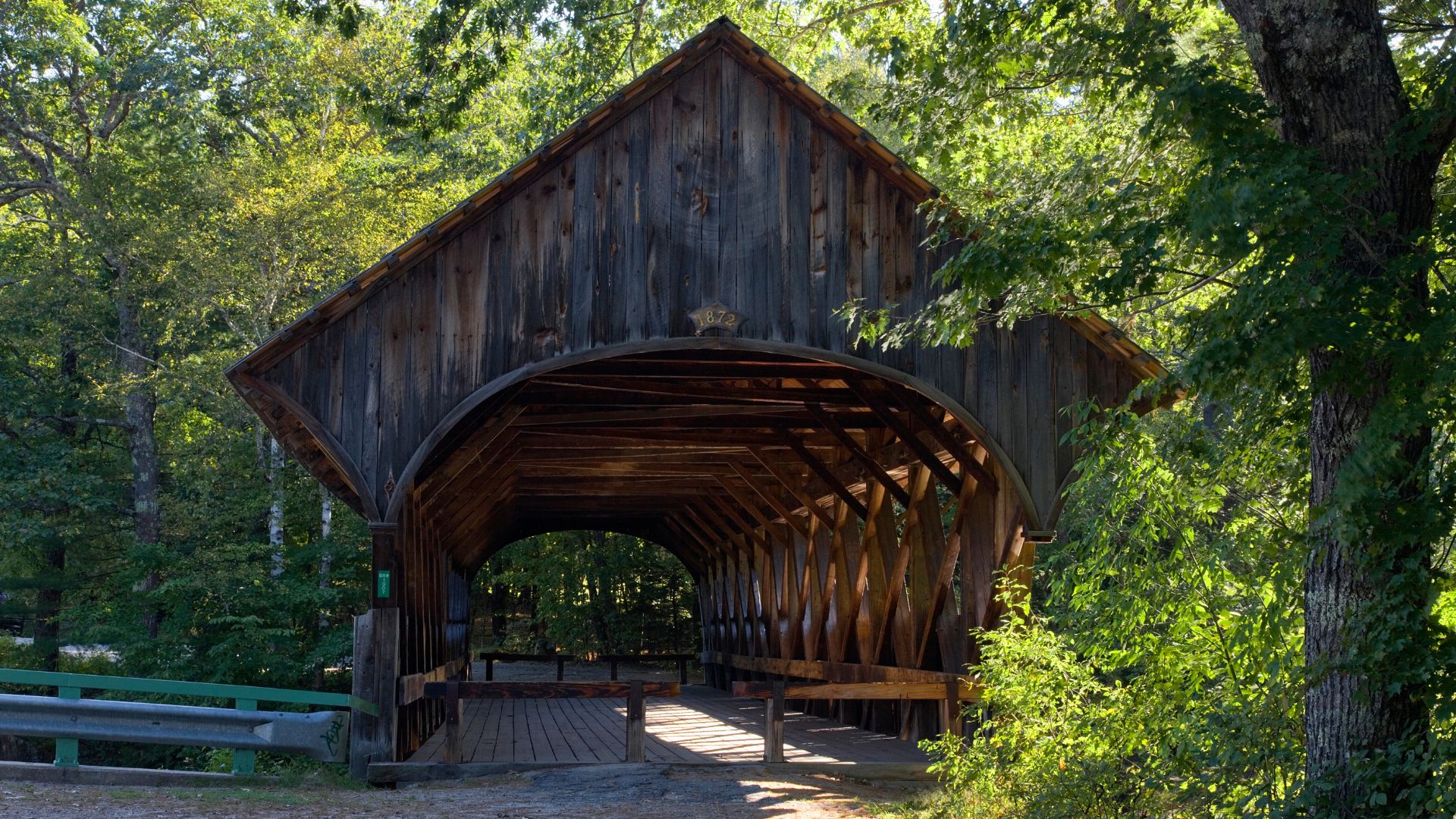 A wooden covered bridge, known as the Artist's Bridge, stands amidst lush green trees under a bright sky. Sunlight streams into the arched entrance of the bridge, illuminating its interior structure. The bridge is surrounded by dense foliage, with a hint of a guardrail visible on the left side, suggesting a road leading to it.