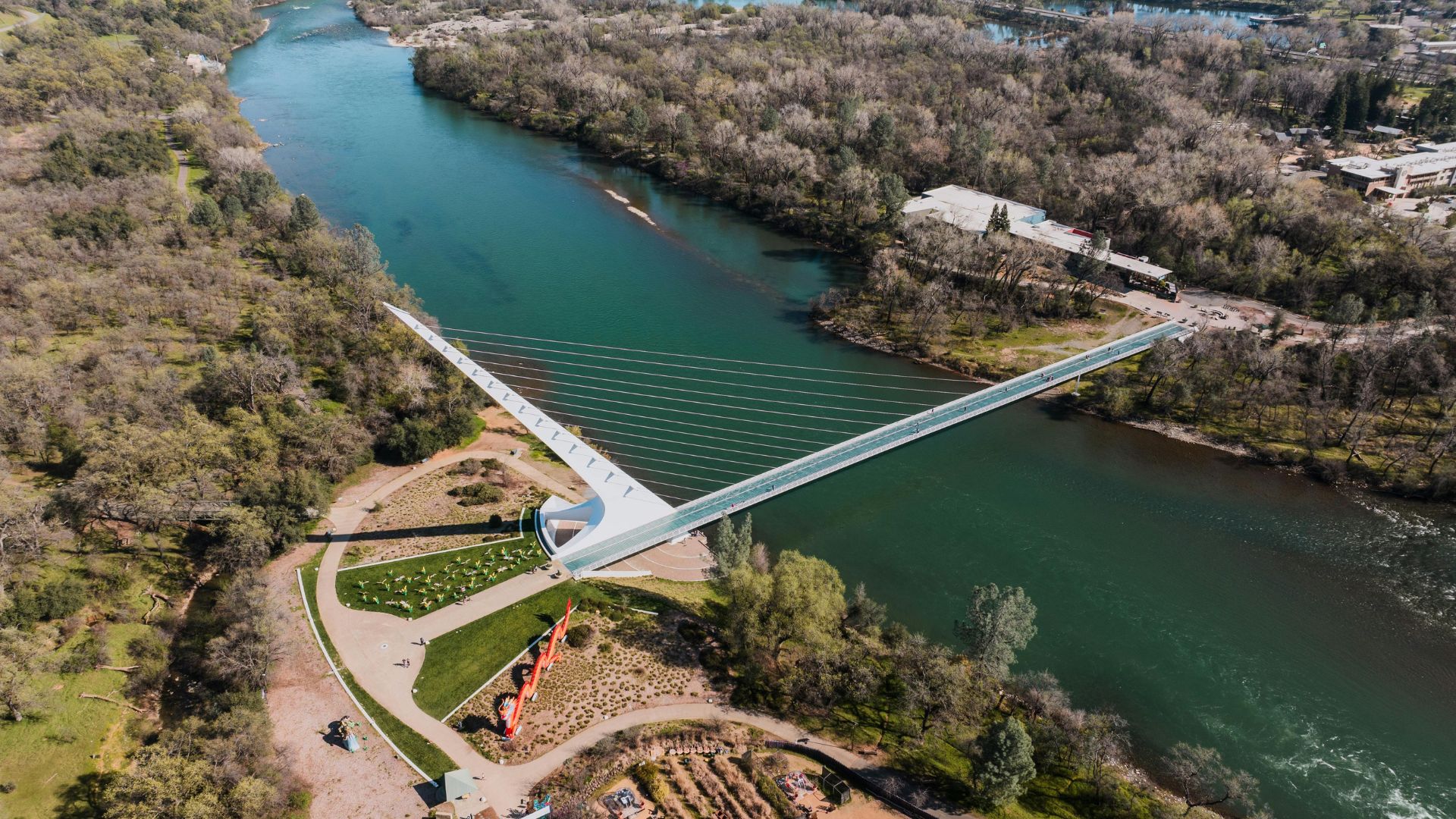 An aerial view of the white, cable-stayed Sundial Bridge spanning a green river, surrounded by lush green and brown trees, with a building visible on the far bank.