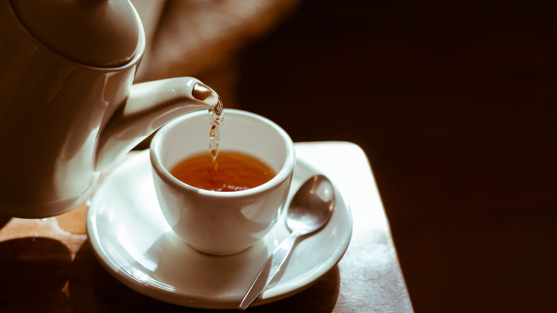 A white teapot pours amber-colored tea into a matching white teacup on a saucer with a spoon, set on a wooden surface.
