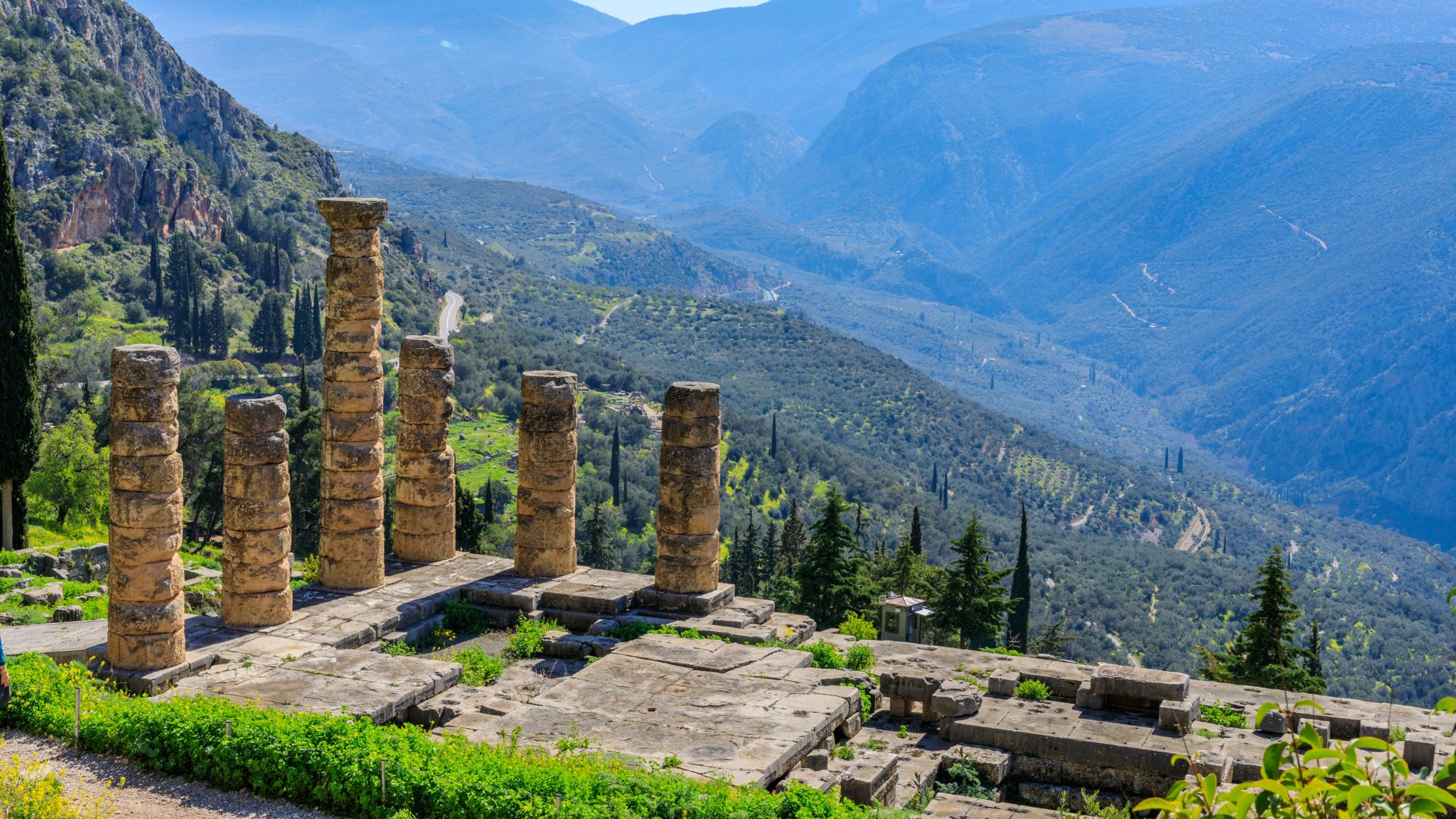 Ruins of the ancient Temple of Apollo in Delphi, Greece, with several standing columns overlooking a vast mountainous landscape under a clear sky.