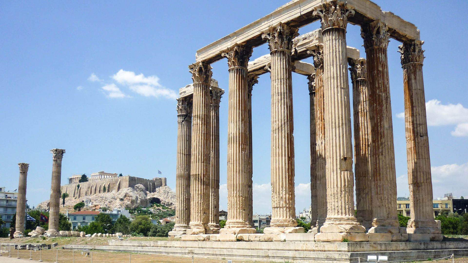 The image shows the ruins of the Temple of Olympian Zeus in Athens, Greece, with several large, fluted Corinthian columns still standing, and the Acropolis visible in the background under a clear blue sky.