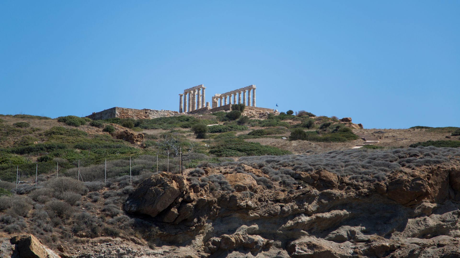 A wide shot of ancient Greek ruins, specifically the white marble columns of the Temple of Poseidon, standing atop a rugged, scrub-covered hill overlooking the sea under a clear blue sky.