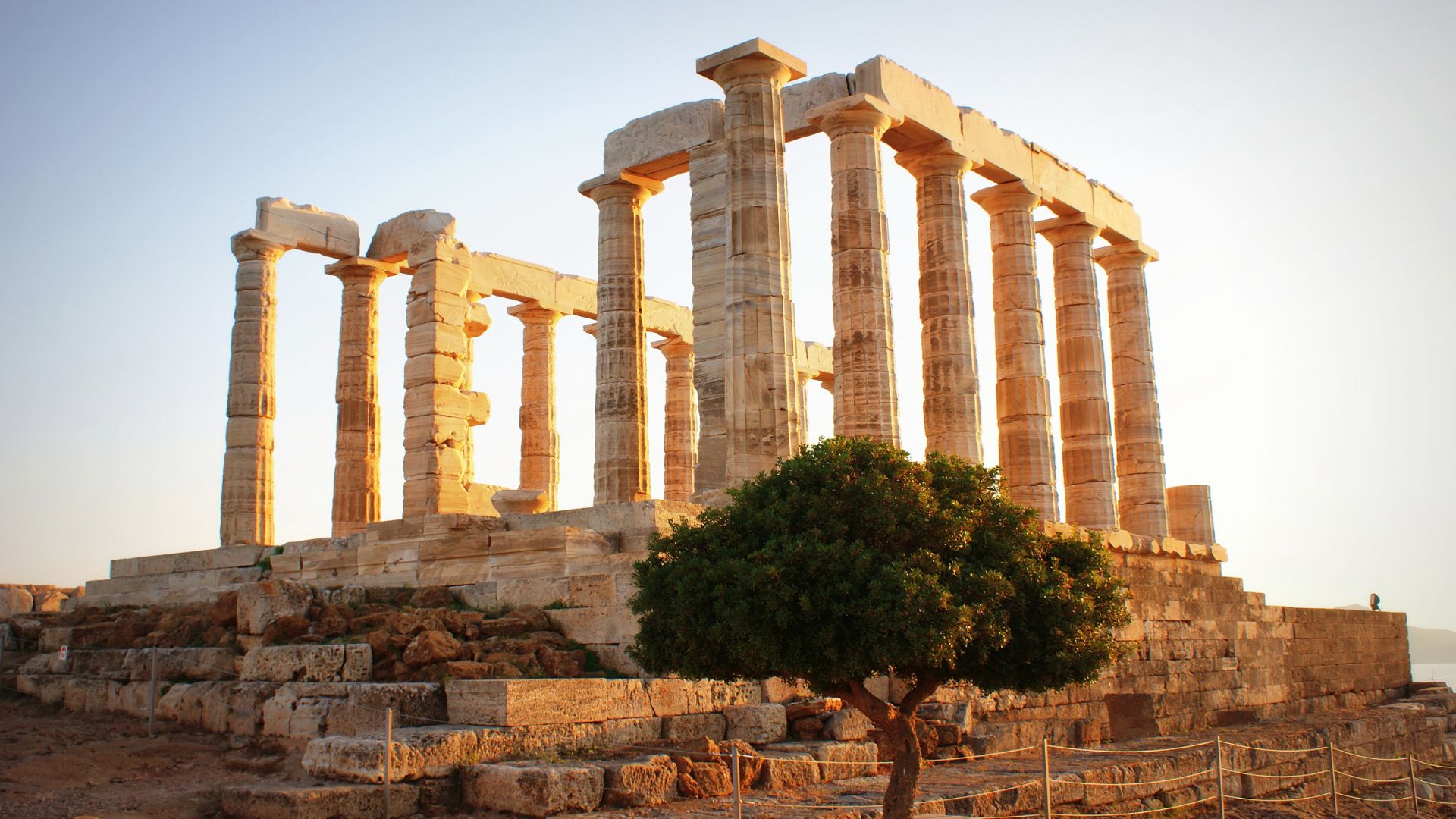 Ancient Greek temple ruins with tall, weathered columns stand on a rocky promontory overlooking a body of water under a clear sky, with a small tree in the foreground.
