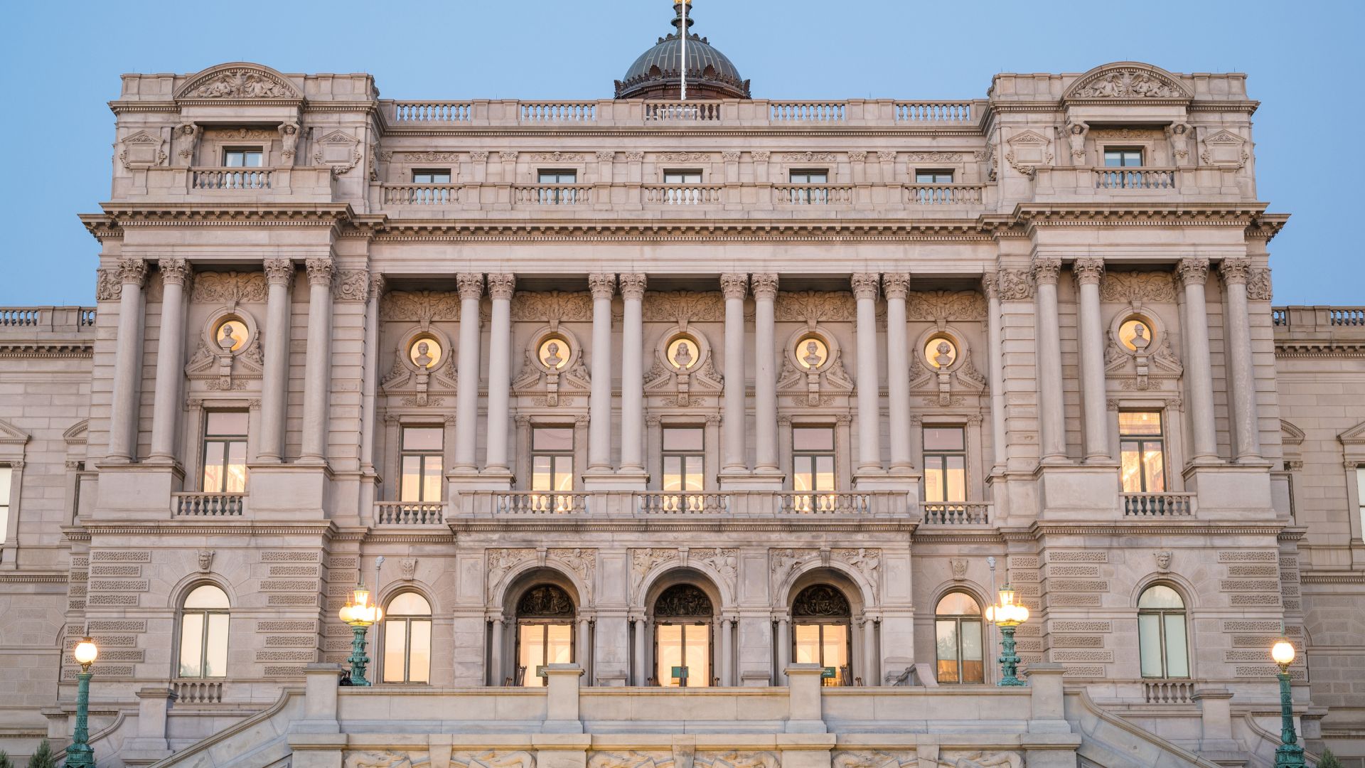 A grand, classical style building with a central dome and numerous arched windows, columns, and intricate architectural details, illuminated by warm interior lights against a twilight sky.