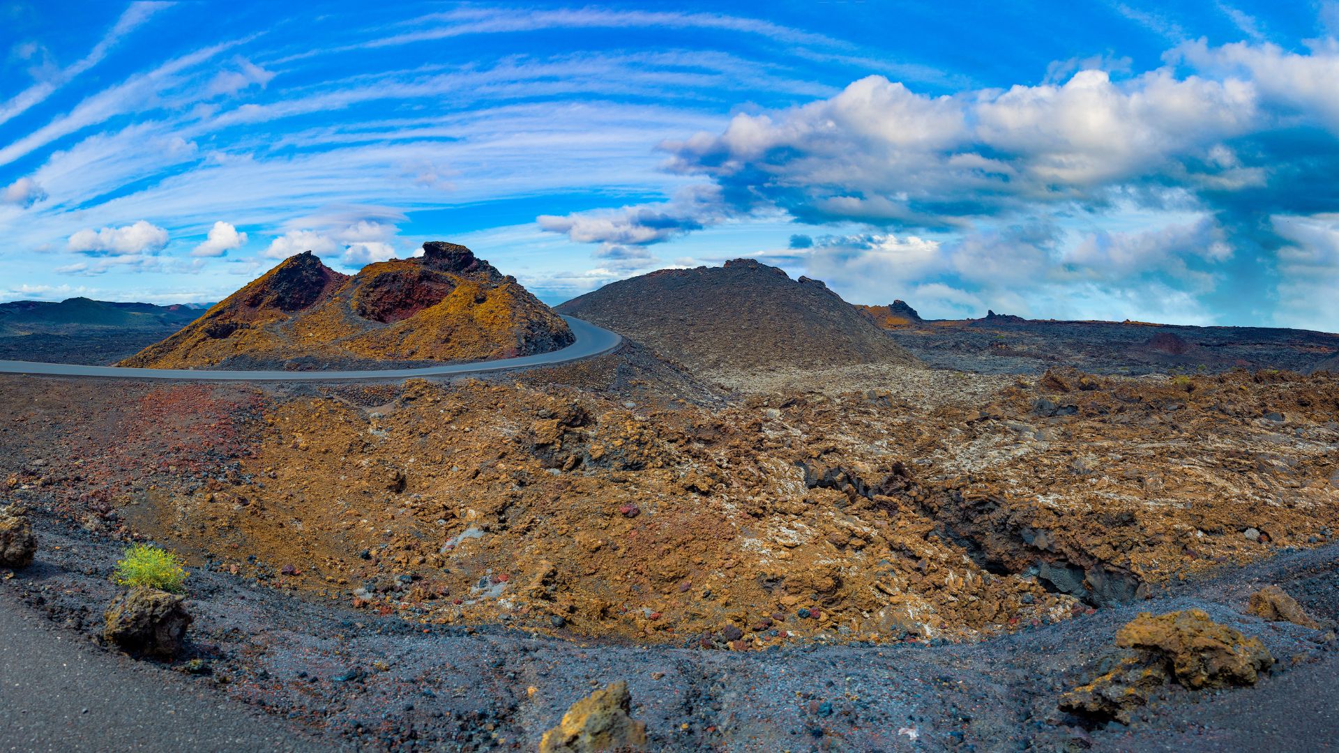 A wide panoramic view of Timanfaya National Park in Lanzarote, showcasing a volcanic landscape with dark, rocky terrain, scattered reddish-brown volcanic cones, and a winding road cutting through the desolate yet striking environment under a blue sky with scattered white clouds.