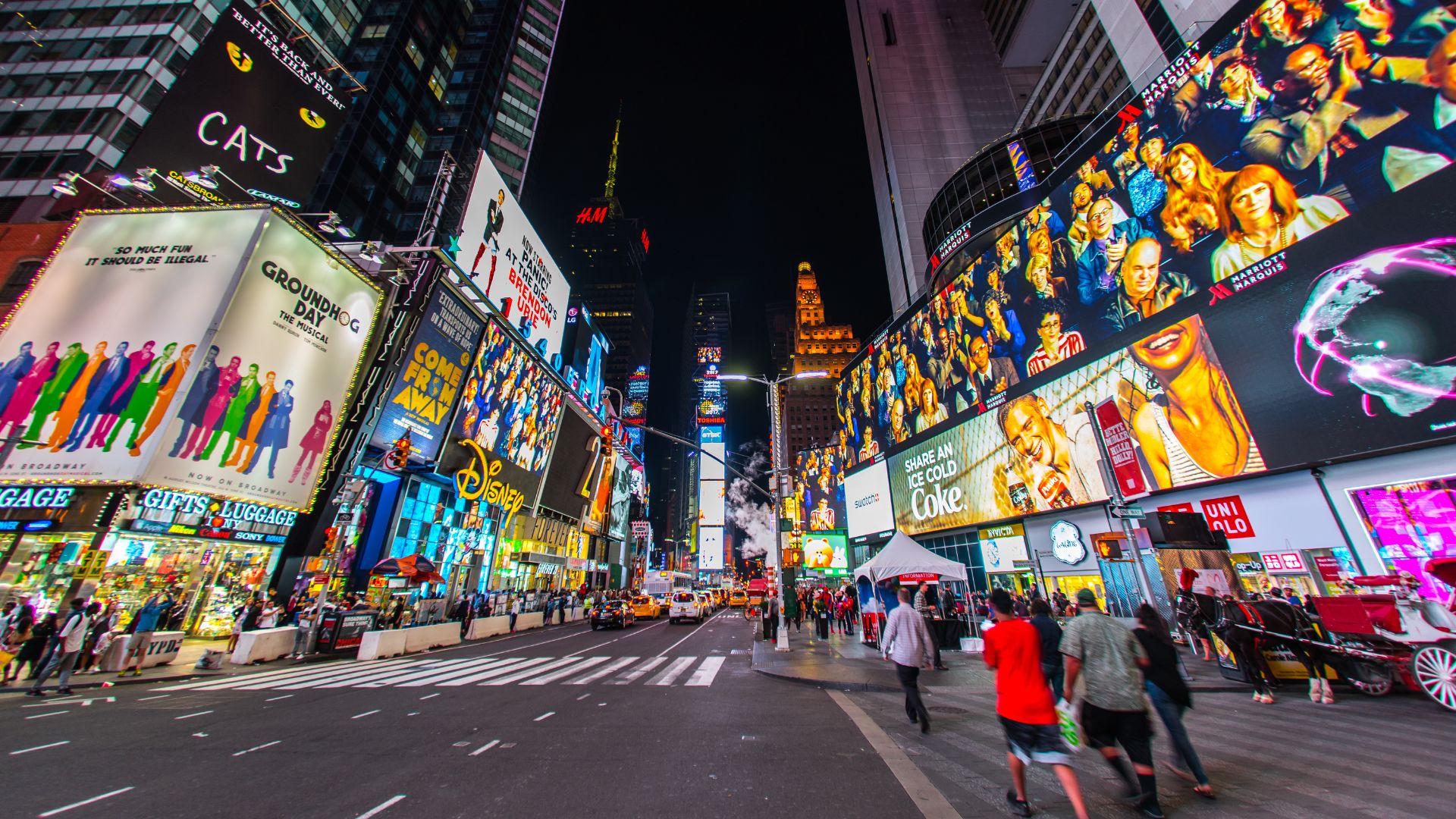 A vibrant, wide-angle night shot of Times Square in New York City, illuminated by numerous large, colorful digital billboards displaying advertisements and entertainment content, with pedestrians walking on the street and sidewalks below. 