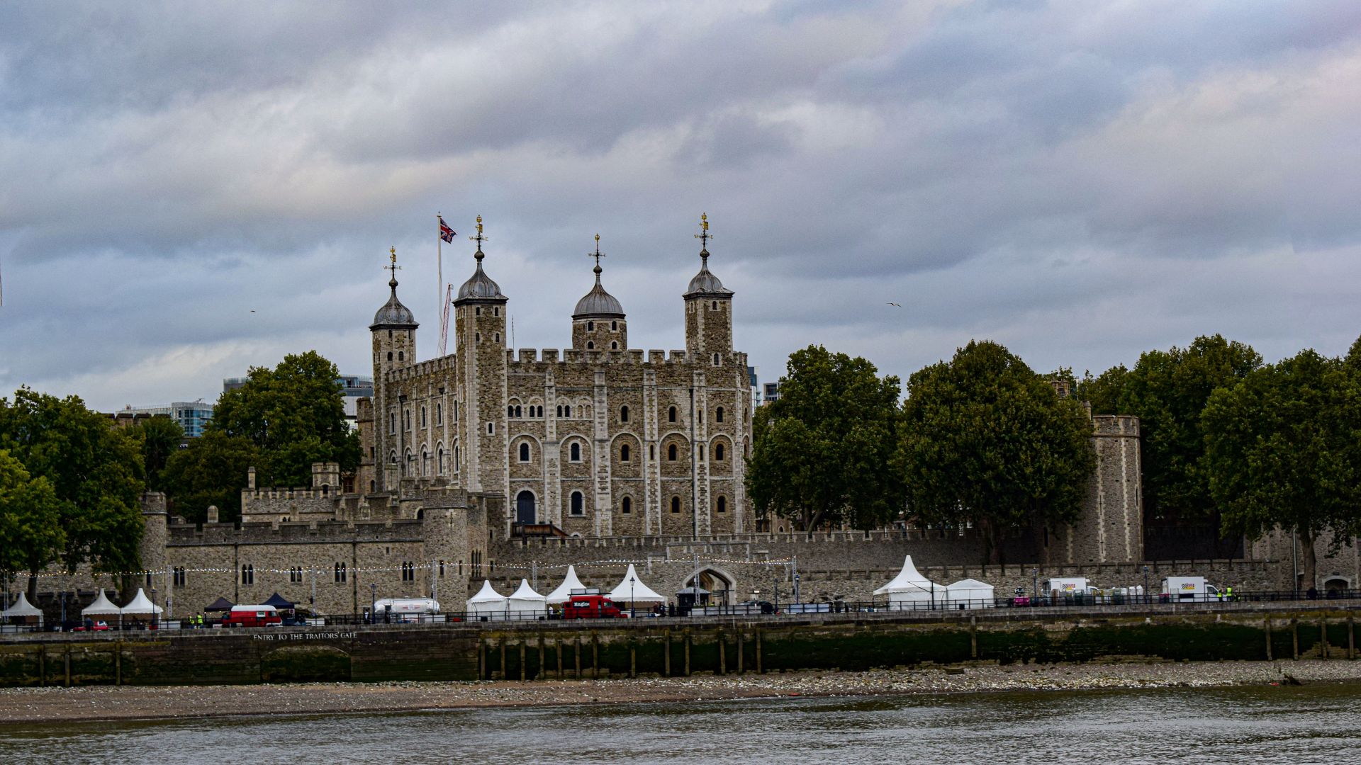A wide shot of the historic Tower of London, a large stone castle with multiple towers and battlements, situated on the bank of a river under a cloudy sky. Green trees line the bank behind the castle, and several white tents are visible along the waterfront in front of the castle walls.