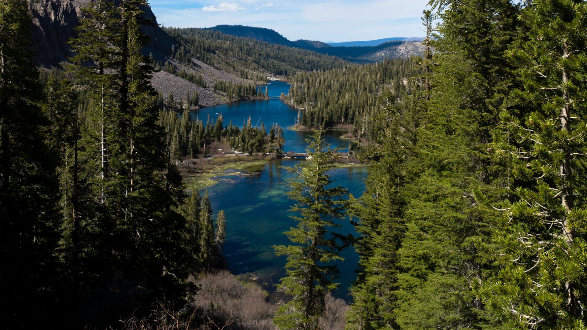 A scenic view of Twin Lakes in California, surrounded by dense evergreen forests and mountains under a clear sky. The two lakes are connected by a narrow passage, with a small bridge visible in the middle ground.
