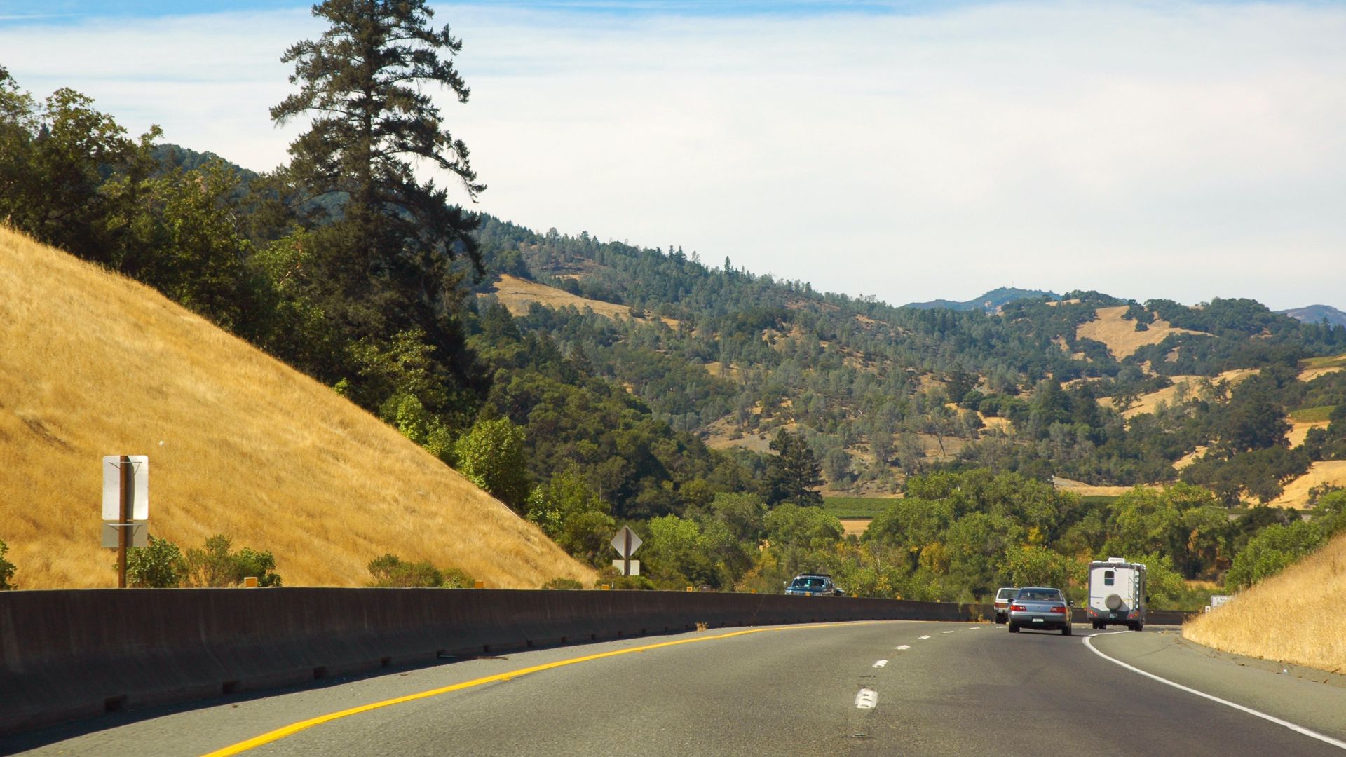 A highway curves through a hilly, golden-grass landscape with scattered trees under a clear sky. Cars are visible on the road, with a large, dry hillside on the left and a more forested, rolling landscape in the distance on the right.