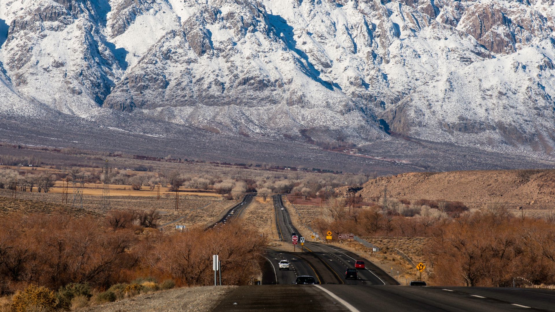 A long, winding highway stretches towards distant, snow-covered mountains under a clear sky, flanked by dry, brush-filled landscapes in the foreground.