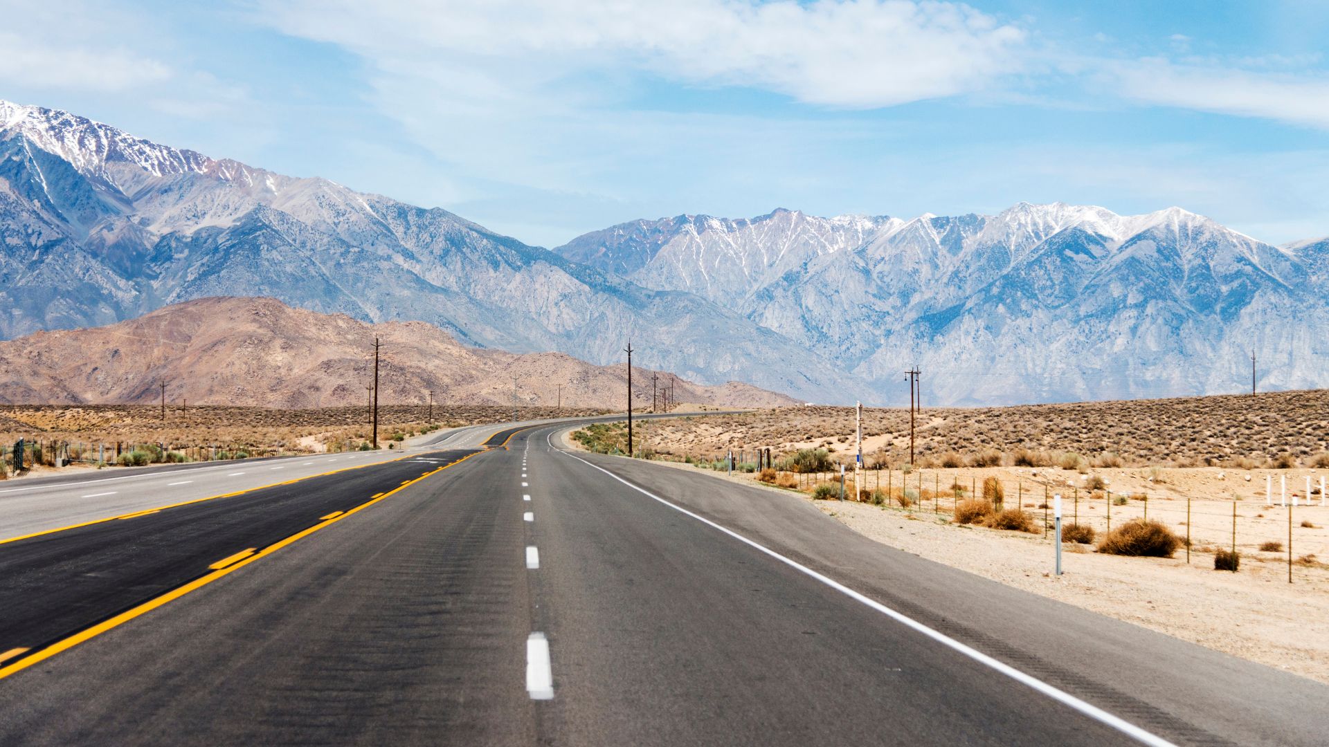 A long, empty highway (U.S. Route 395) stretches towards a range of majestic, snow-capped mountains under a clear sky in the Eastern Sierra region of California.