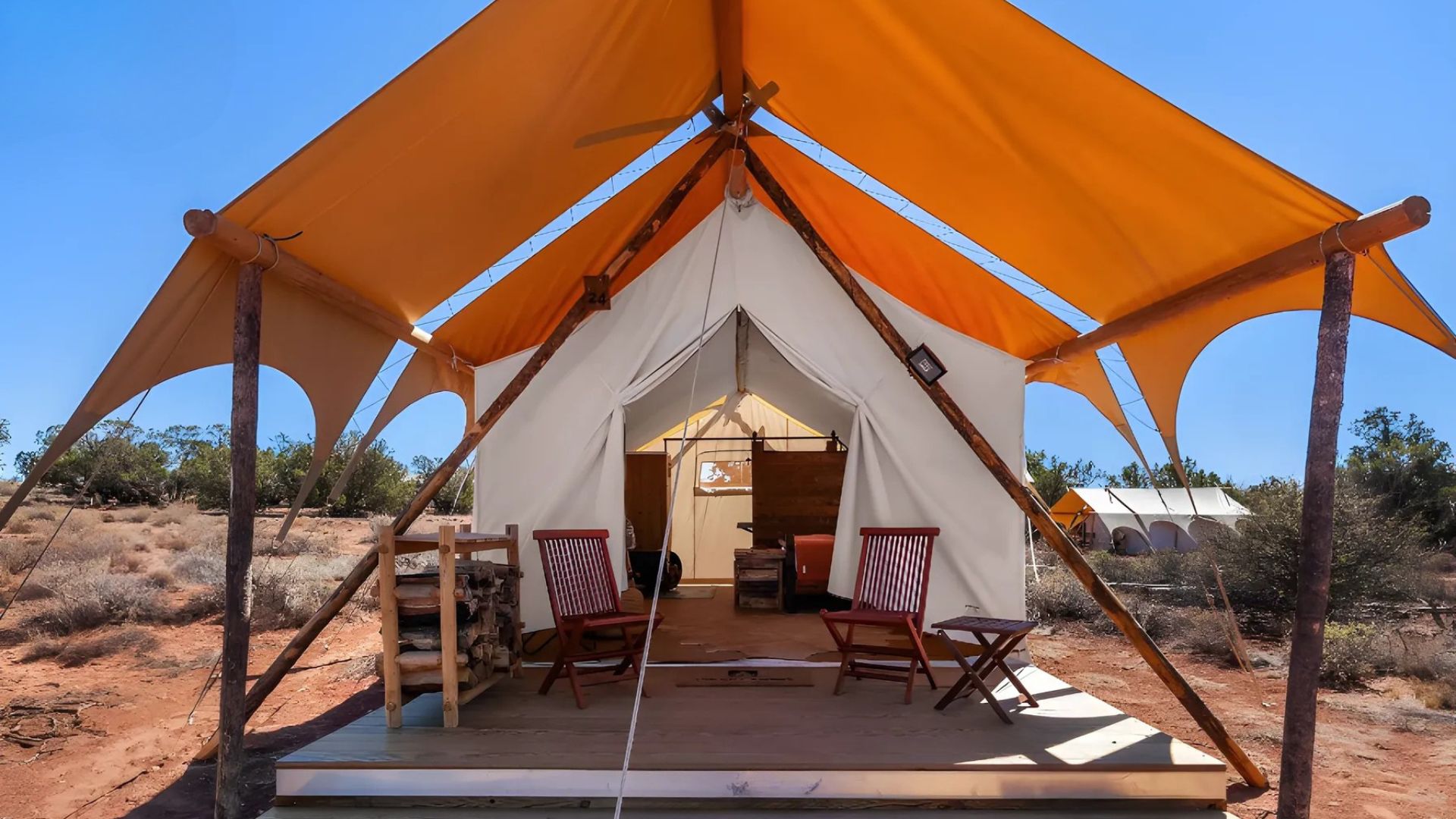 A large, luxurious safari-style tent with an orange overhead canopy and wooden deck sits in a sunny, arid landscape, furnished with two red chairs and a small table on the deck.