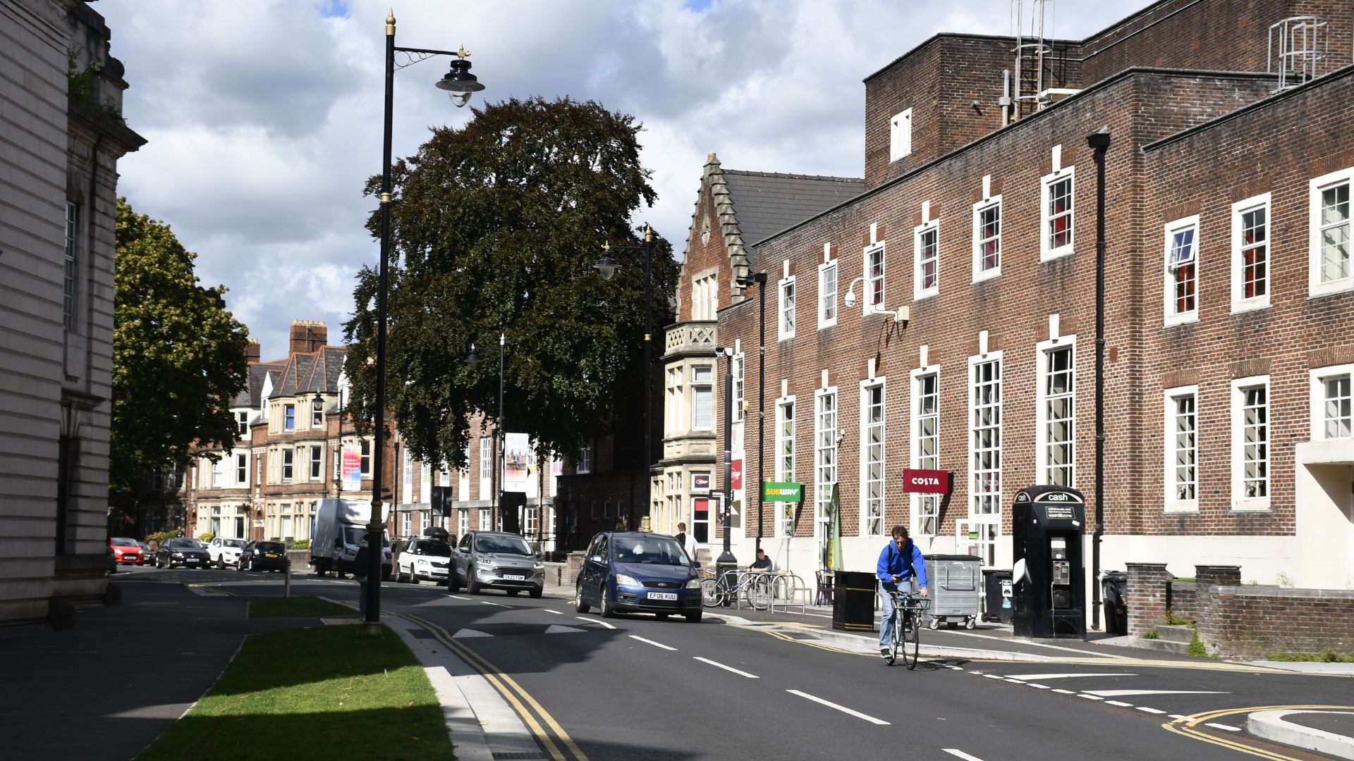 A street view of Tunbridge Wells Library, a red-brick building with multiple windows, on a cloudy day with cars on the road and a person on a bicycle in the foreground.