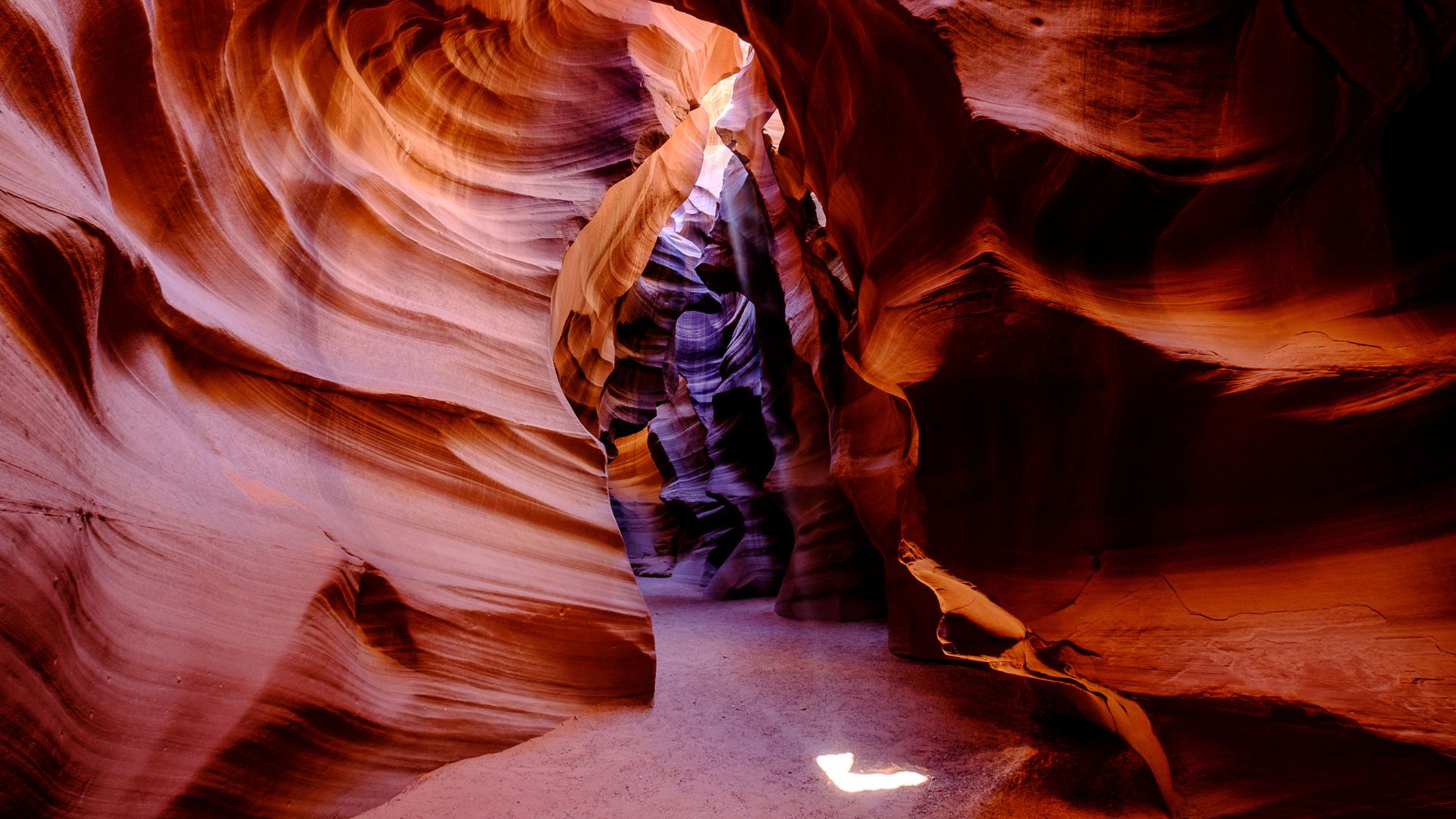 A narrow slot canyon with swirling, orange-red sandstone walls, illuminated by shafts of light filtering down from above, revealing the smooth, sculpted textures of the rock formations.