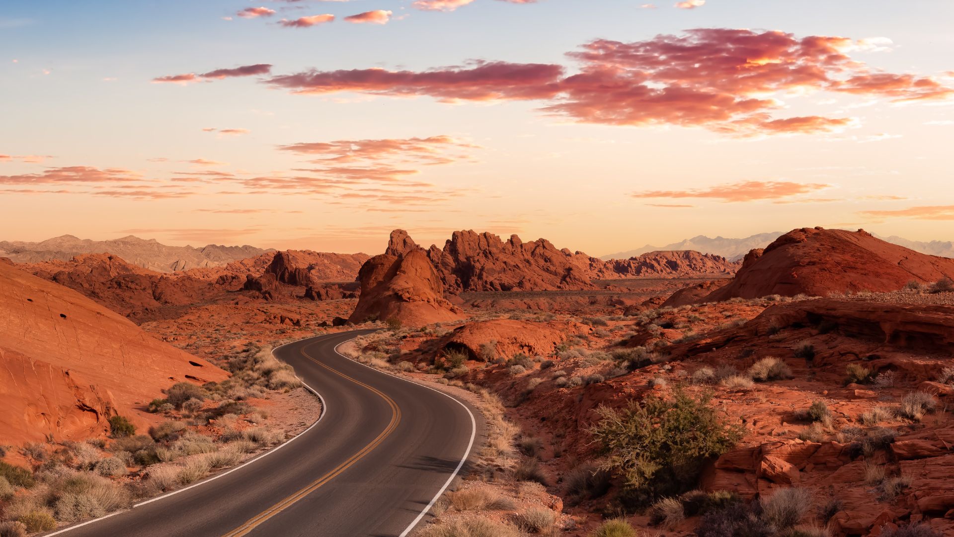 A winding road curves through a vibrant red rock desert landscape under a colorful sunset sky, with various rock formations and sparse desert vegetation visible.