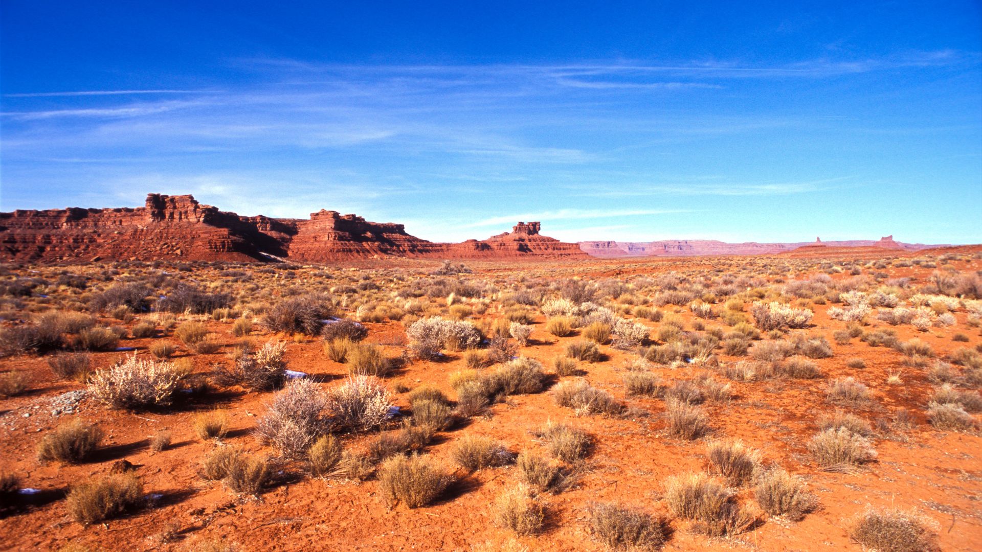 A wide-angle view of the Valley of the Gods in Utah, showcasing a vast, arid landscape with reddish-orange dirt, scattered desert shrubs, and towering red sandstone mesas and buttes under a clear blue sky.