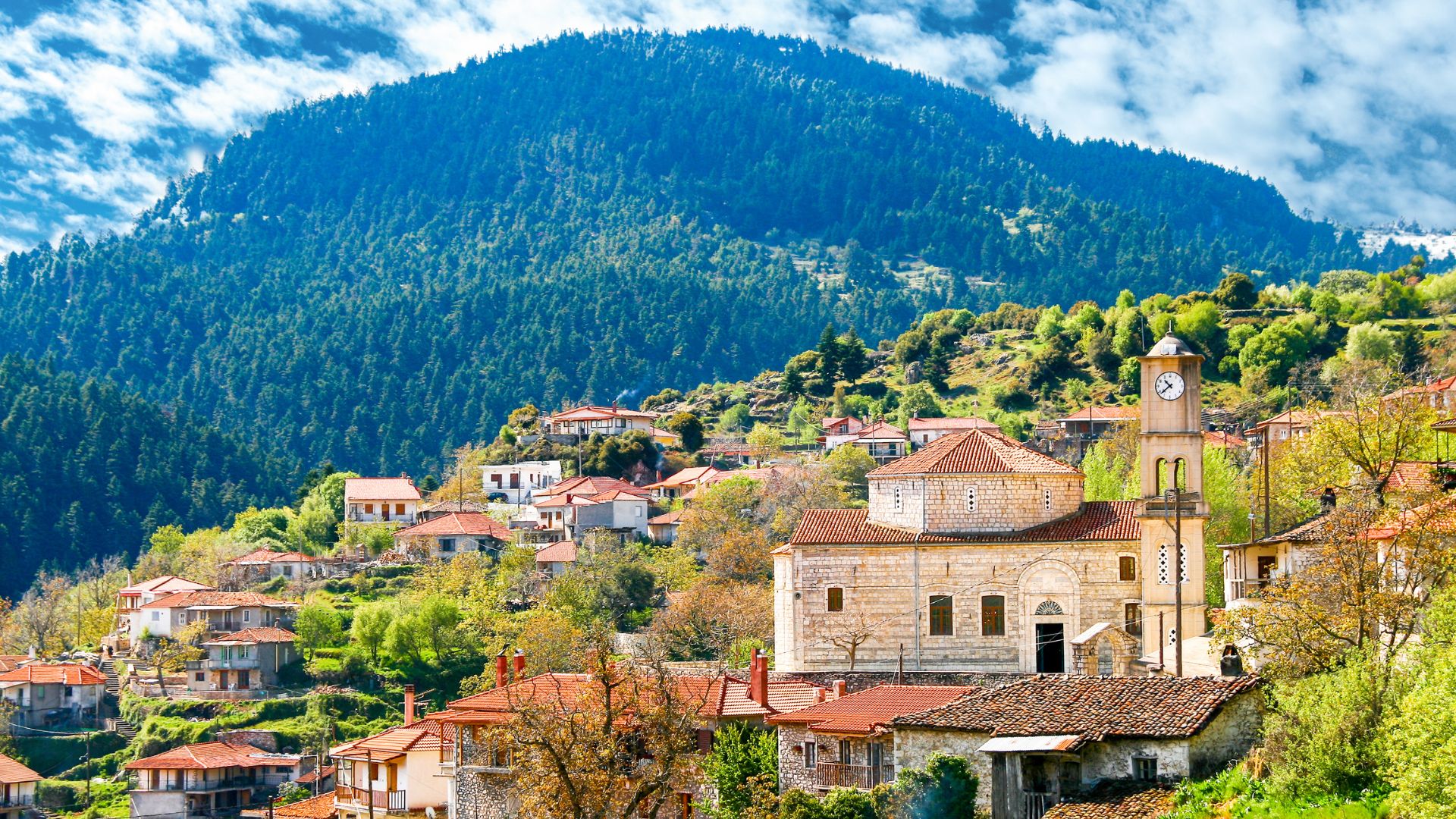 A picturesque mountain village in Greece, Valtesiniko, featuring traditional stone houses with red-tiled roofs, a prominent church with a clock tower, nestled on a verdant hillside with a large, forested mountain in the background under a cloudy sky.