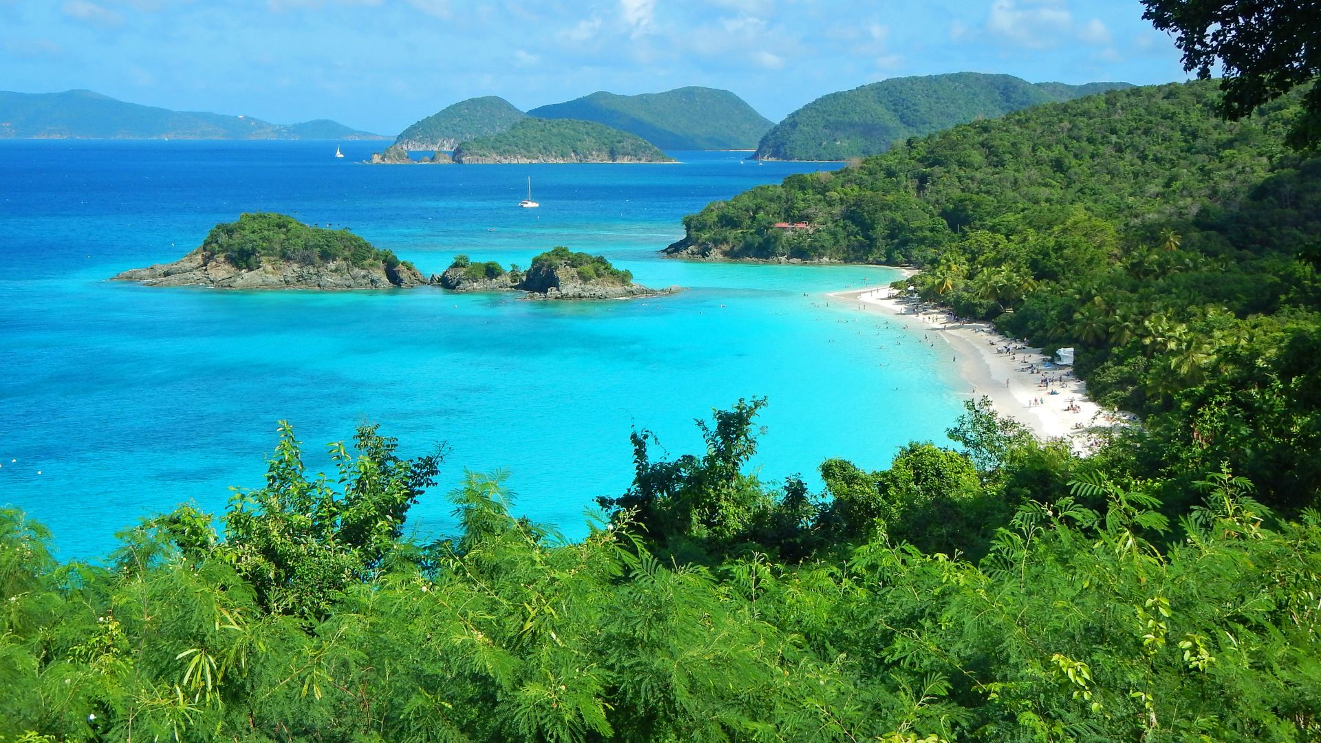 A panoramic view of Trunk Bay in St. John, U.S. Virgin Islands, featuring a white sand beach, vibrant turquoise waters, lush green hills, and small islands dotting the bay, all under a clear blue sky.