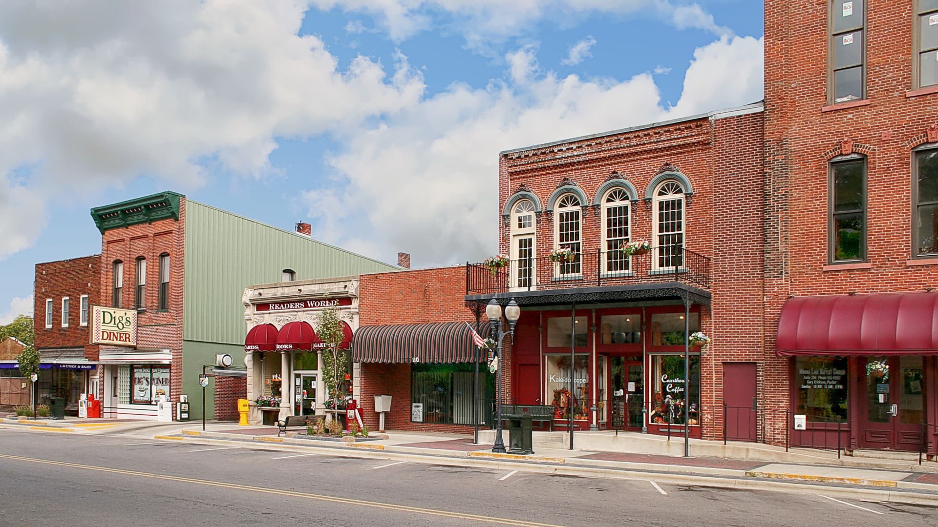 A panoramic view of a quaint downtown street in Warsaw, Indiana, featuring brick buildings with various architectural styles, including a prominent building with arched windows and a balcony, and a green-sided building housing "Digis Diner" on the left. A street with parked cars and a clear sky with scattered clouds are also visible.